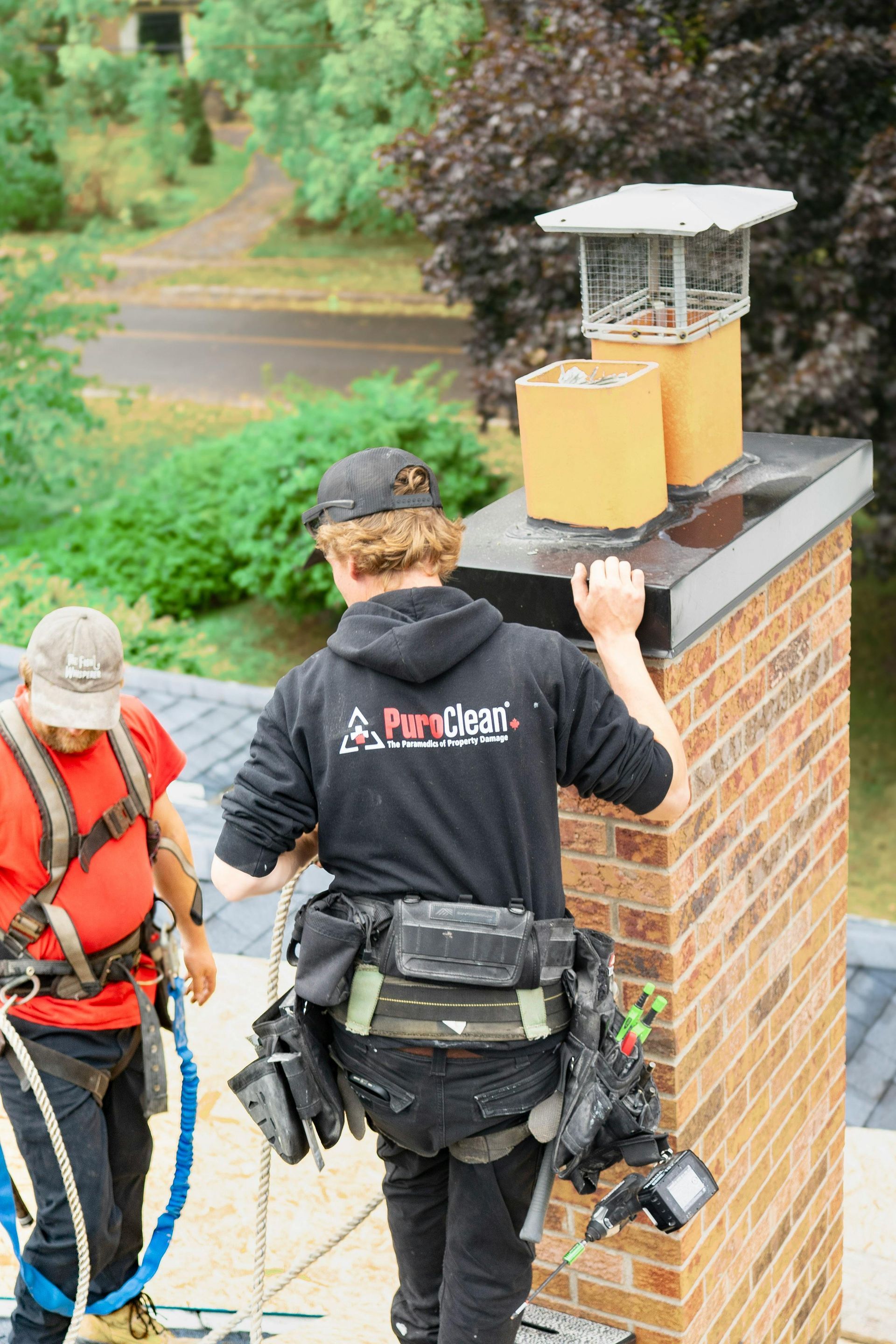 Two workers on a rooftop, one on a chimney, wearing safety harnesses.
