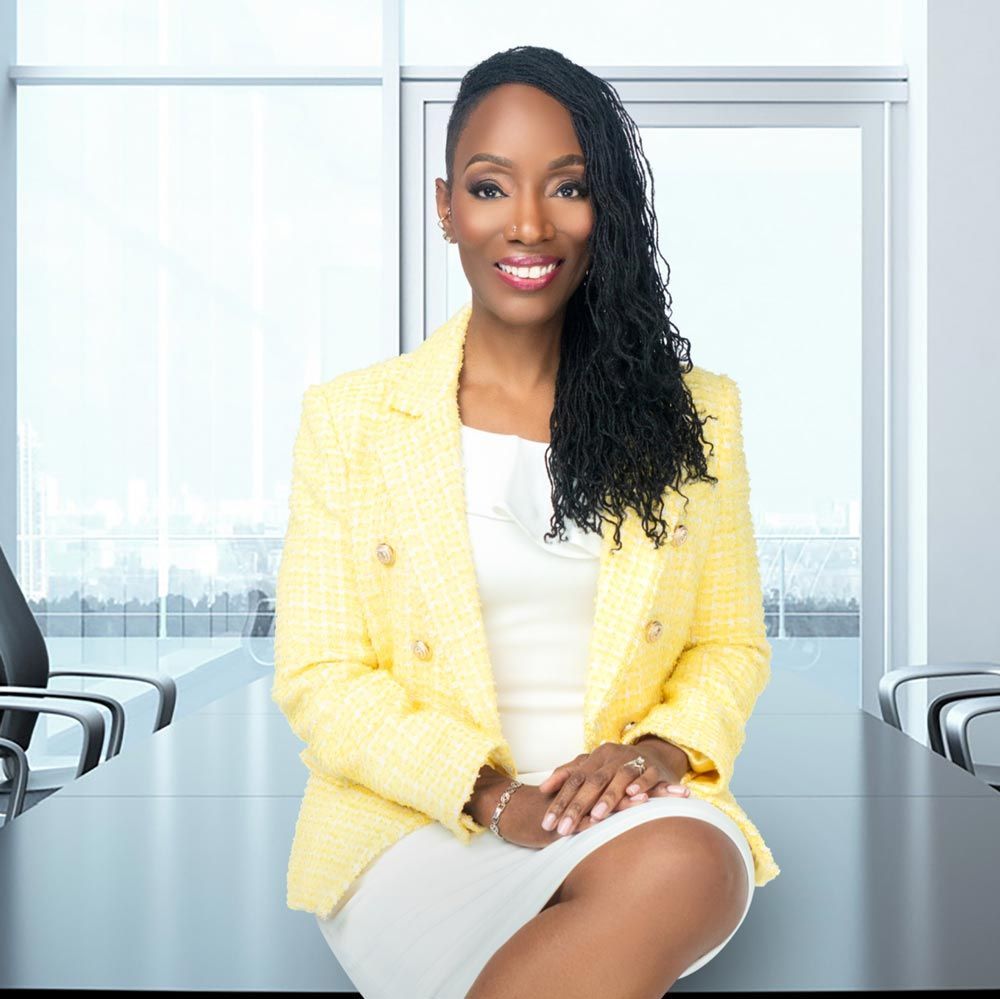 A woman in a yellow jacket and white dress is sitting at a conference table.
