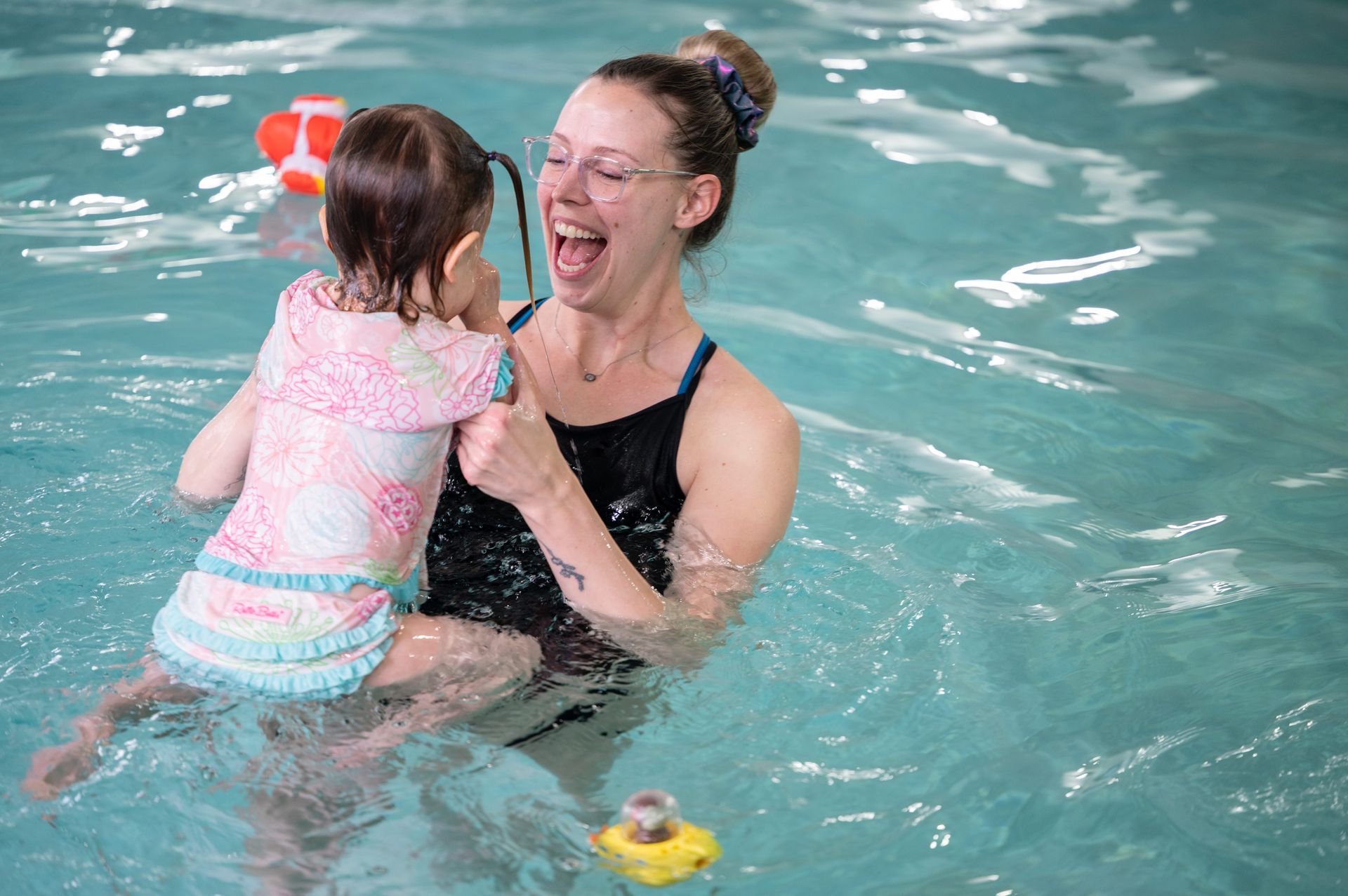 Woman in a pool holds a small child, both laughing. The woman is wearing a swimsuit, the child, a swim top.