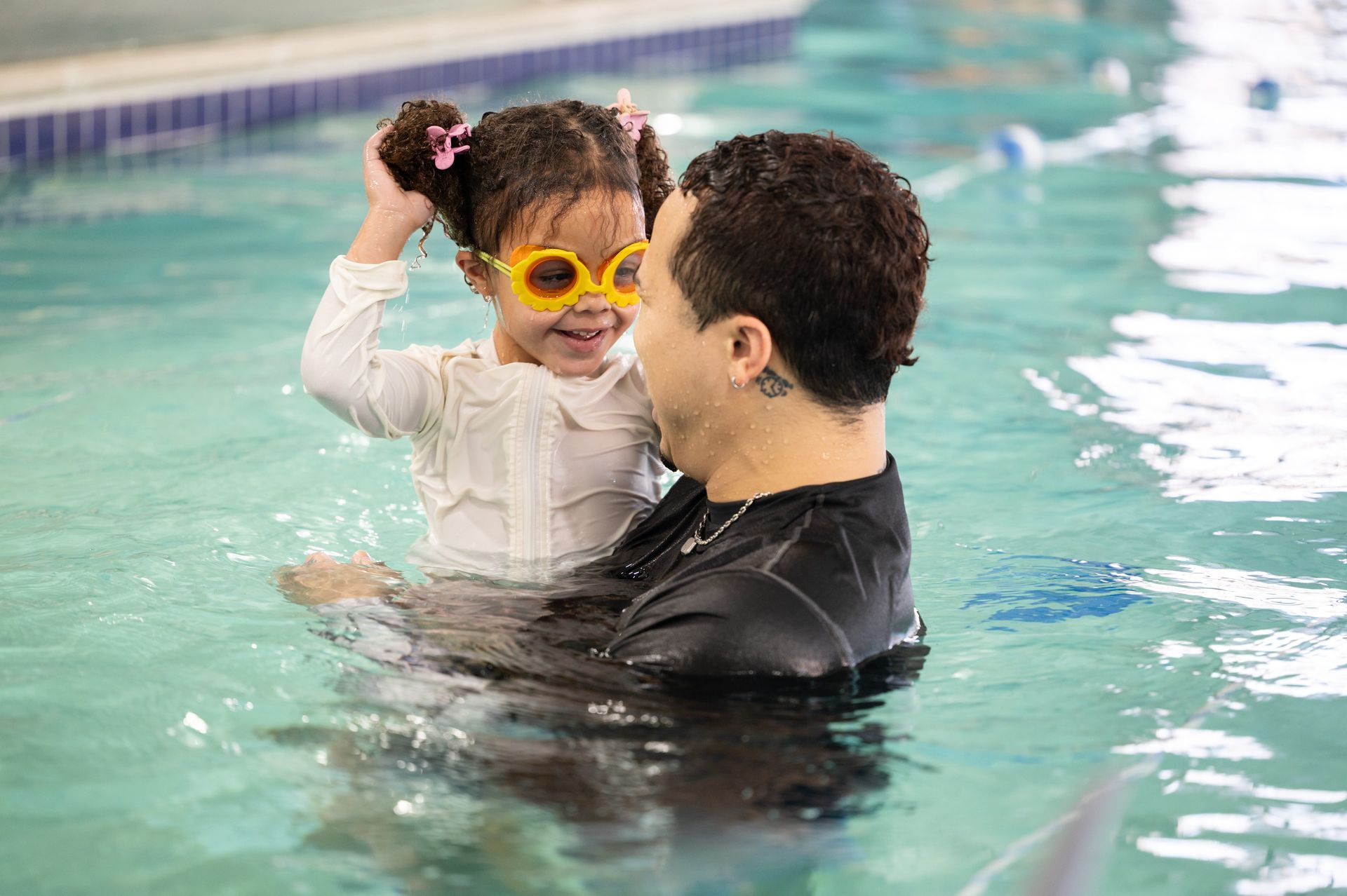 Man holding smiling child in a pool, both wet; child wears yellow sunglasses.