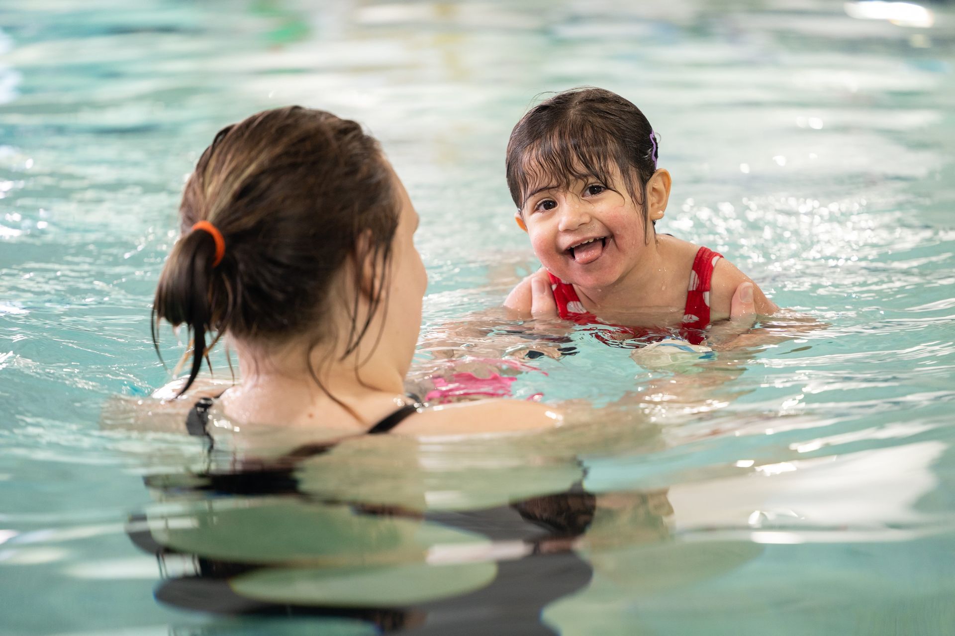 Woman and child in a pool, the child smiling, wearing a red swimsuit, and the woman with an orange hair tie.