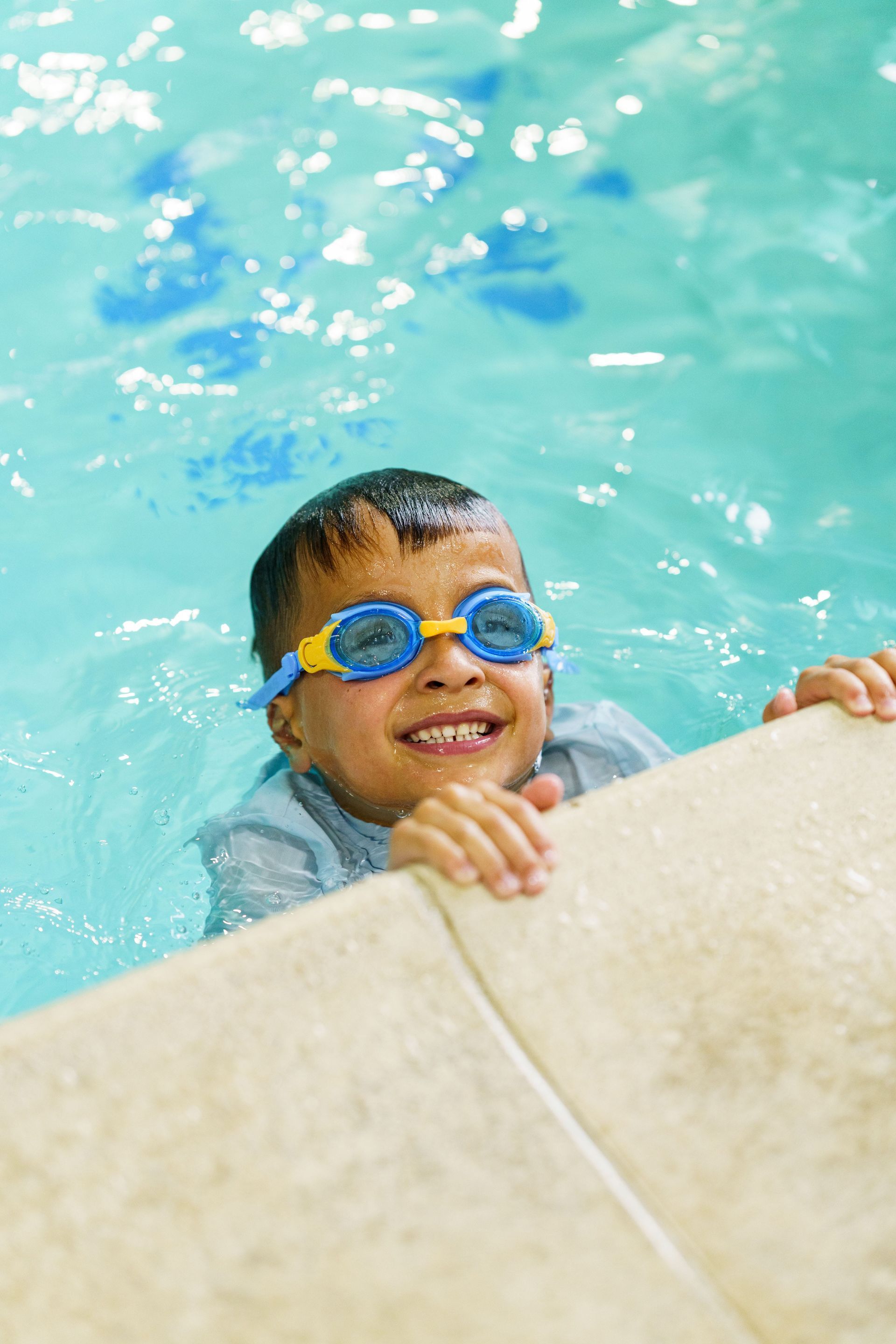 Boy in blue swim goggles smiles while holding the pool edge, submerged in water.