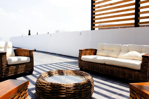 Wicker patio furniture with cream cushions and a round glass-top table on a sunny rooftop terrace with a wooden pergola.