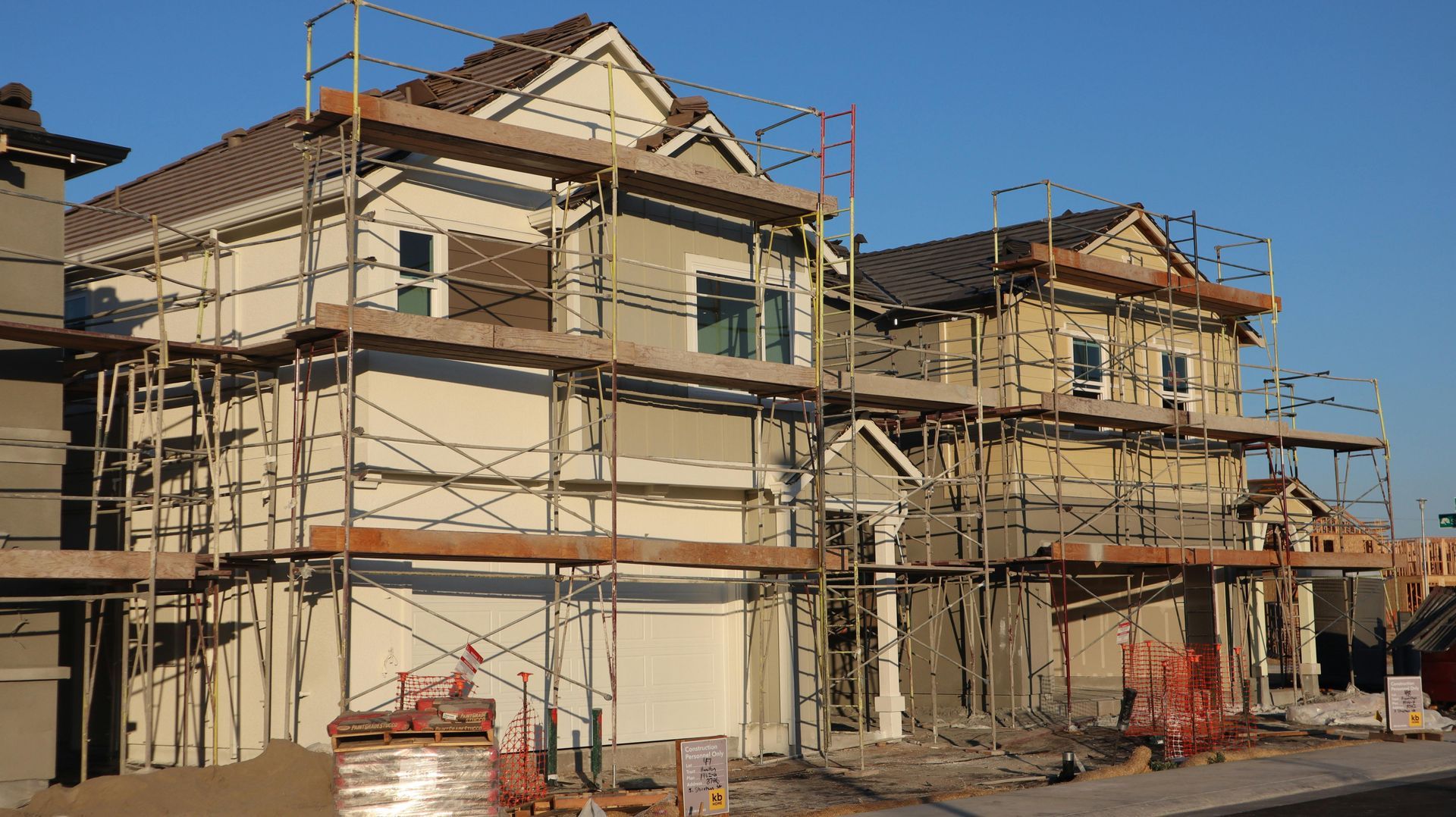 Two houses under construction with metal scaffolding surrounding their light-colored stucco exteriors under a blue sky.