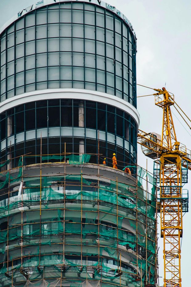 Construction workers on scaffolding around a partially finished, cylindrical glass building with a yellow crane nearby.