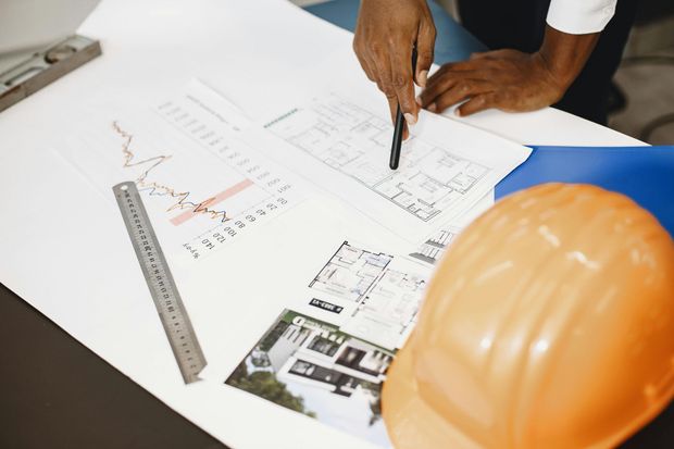 An orange hard hat sits beside architectural blueprints and a ruler on a desk, with hands pointing to the plans.