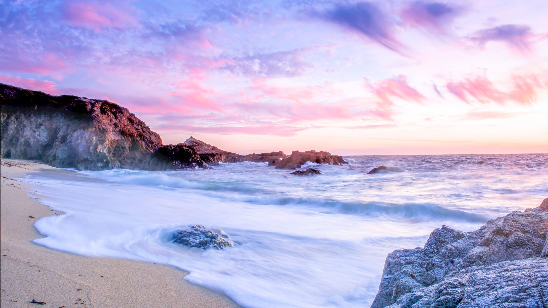 foamy ocean surf coming up onto a sandy beach between rocky outcrops with a blue and pink cloudy sun