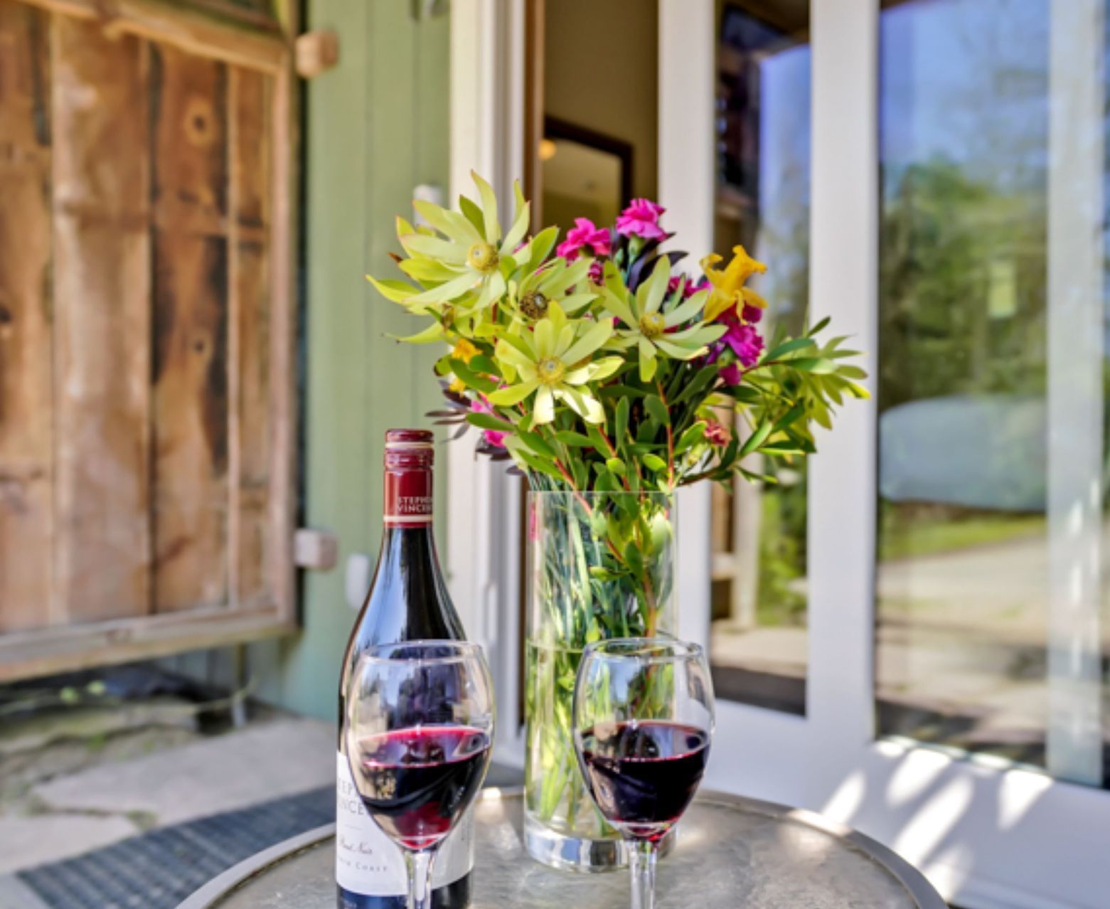 A bottle of wine sits on a table next to two glasses and a vase of flowers