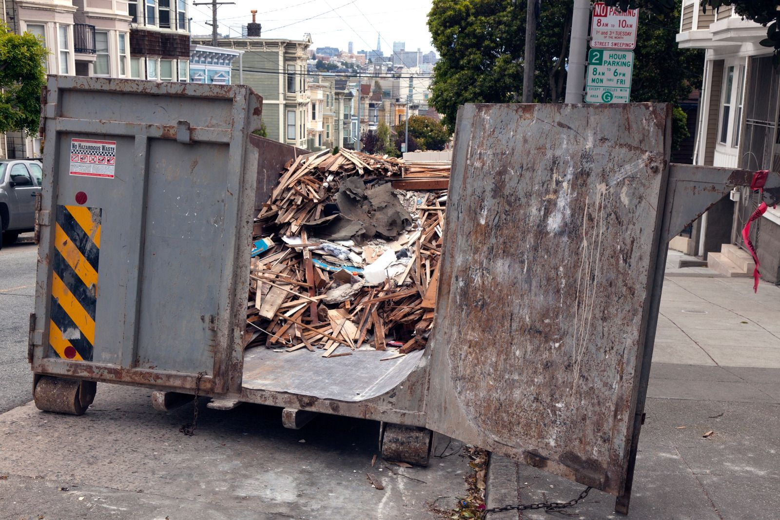 Gray dumpster filled with debris on a city street, partially blocking the sidewalk.