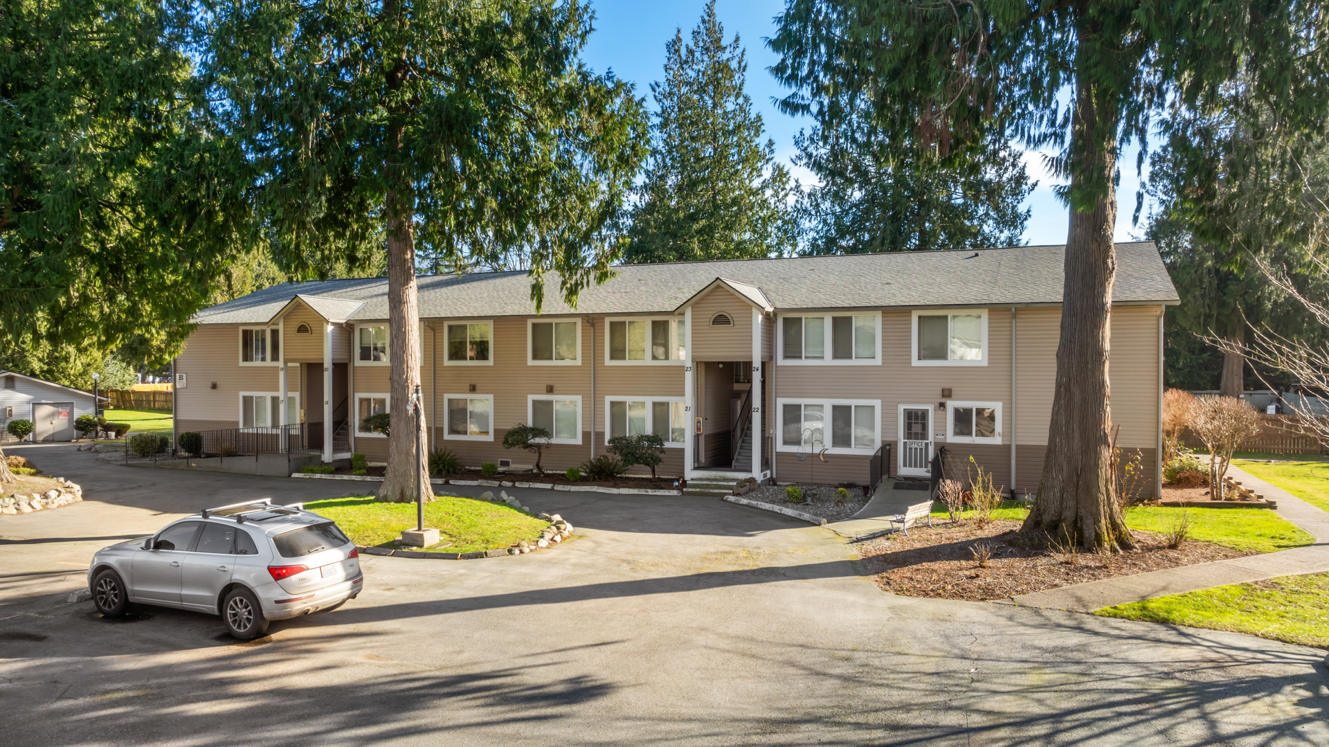 Multi-unit apartment building with parked car in the driveway, trees and grass surrounding.