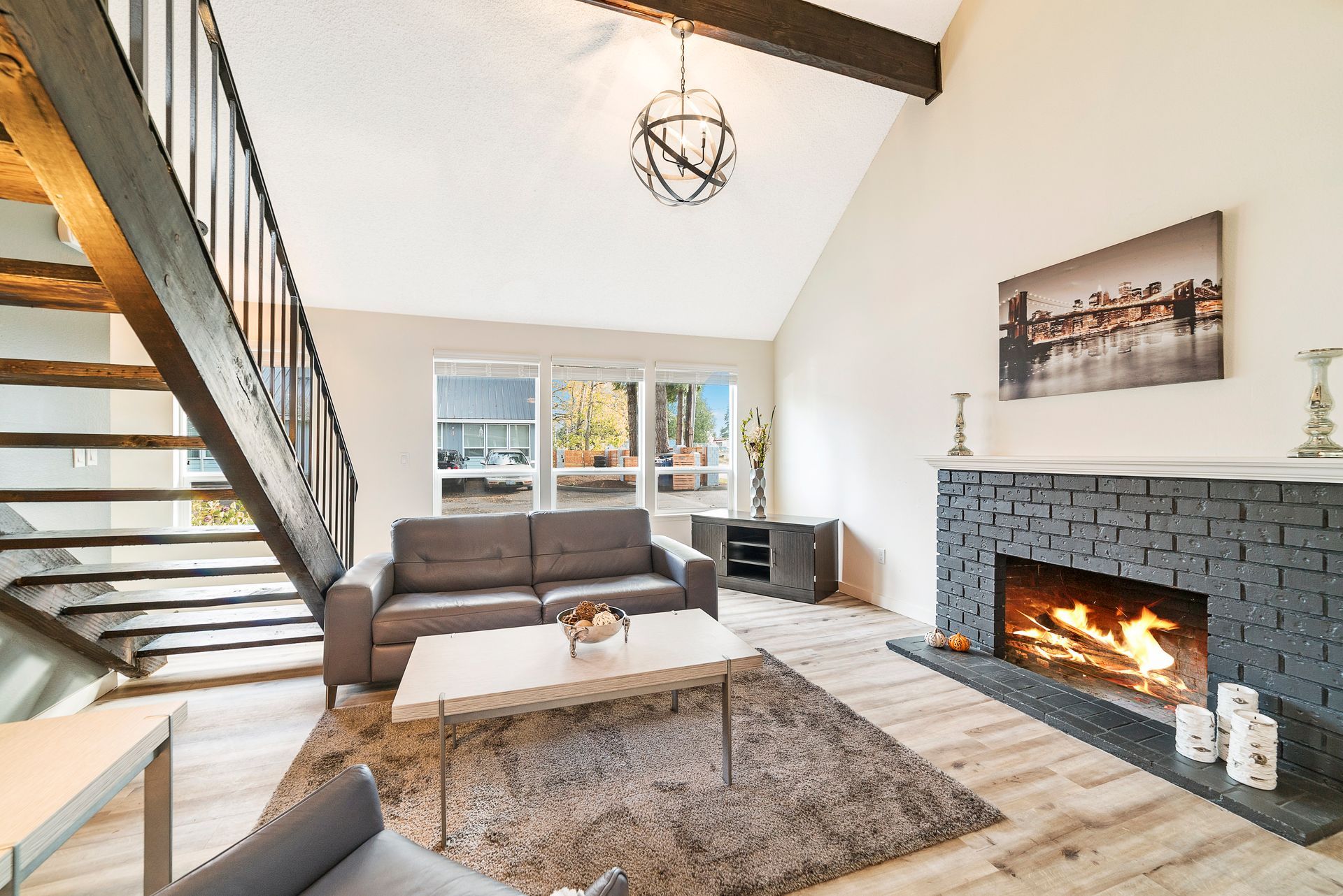 Living room with fireplace, gray sofa, coffee table, wood stairs, and large windows.