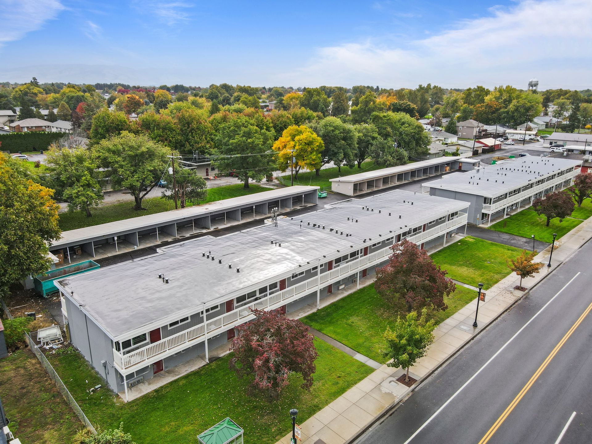 Aerial view of a gray apartment complex with flat roofs, trees, and a road.