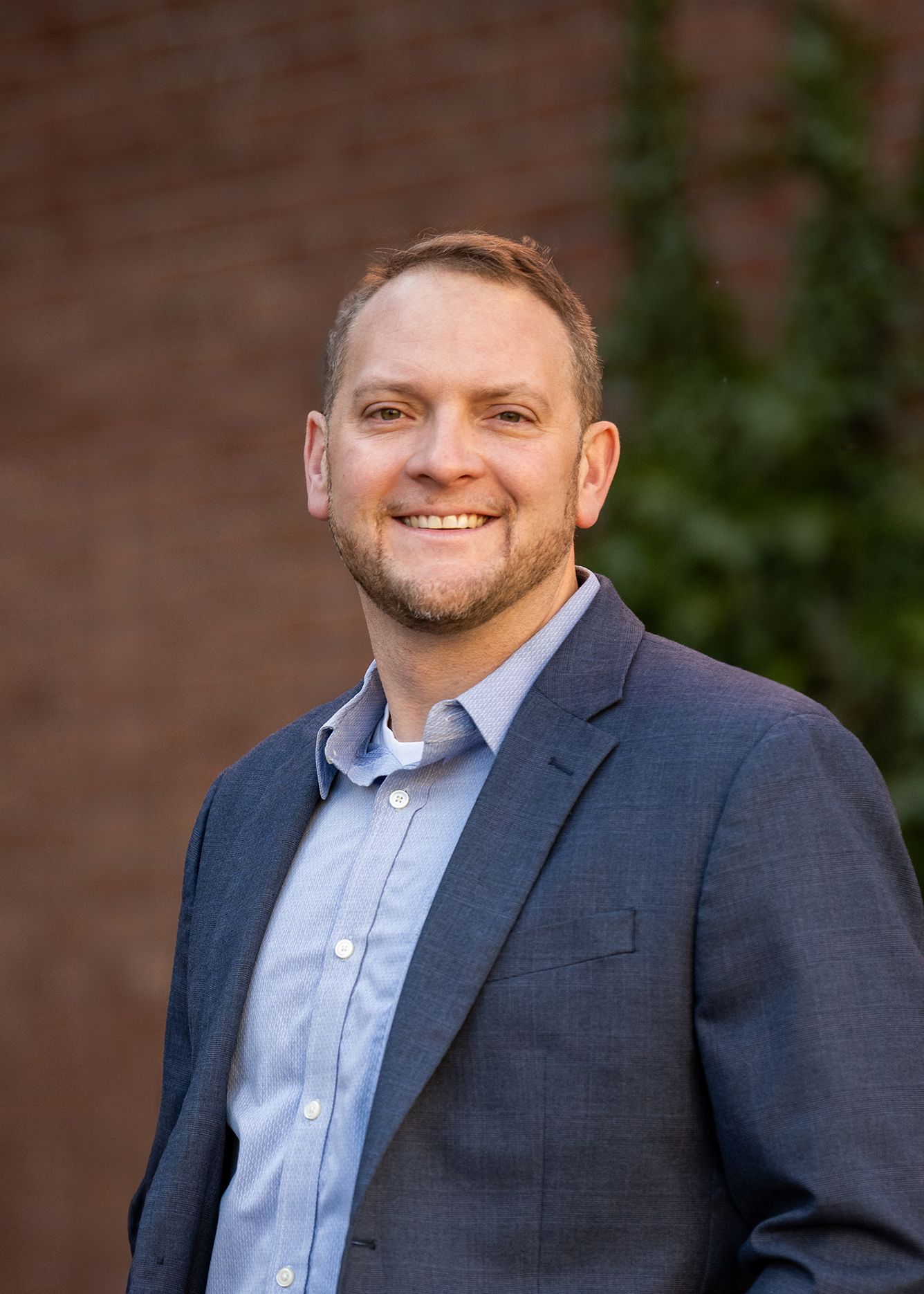 A man in a suit and blue shirt is smiling in front of a brick wall.