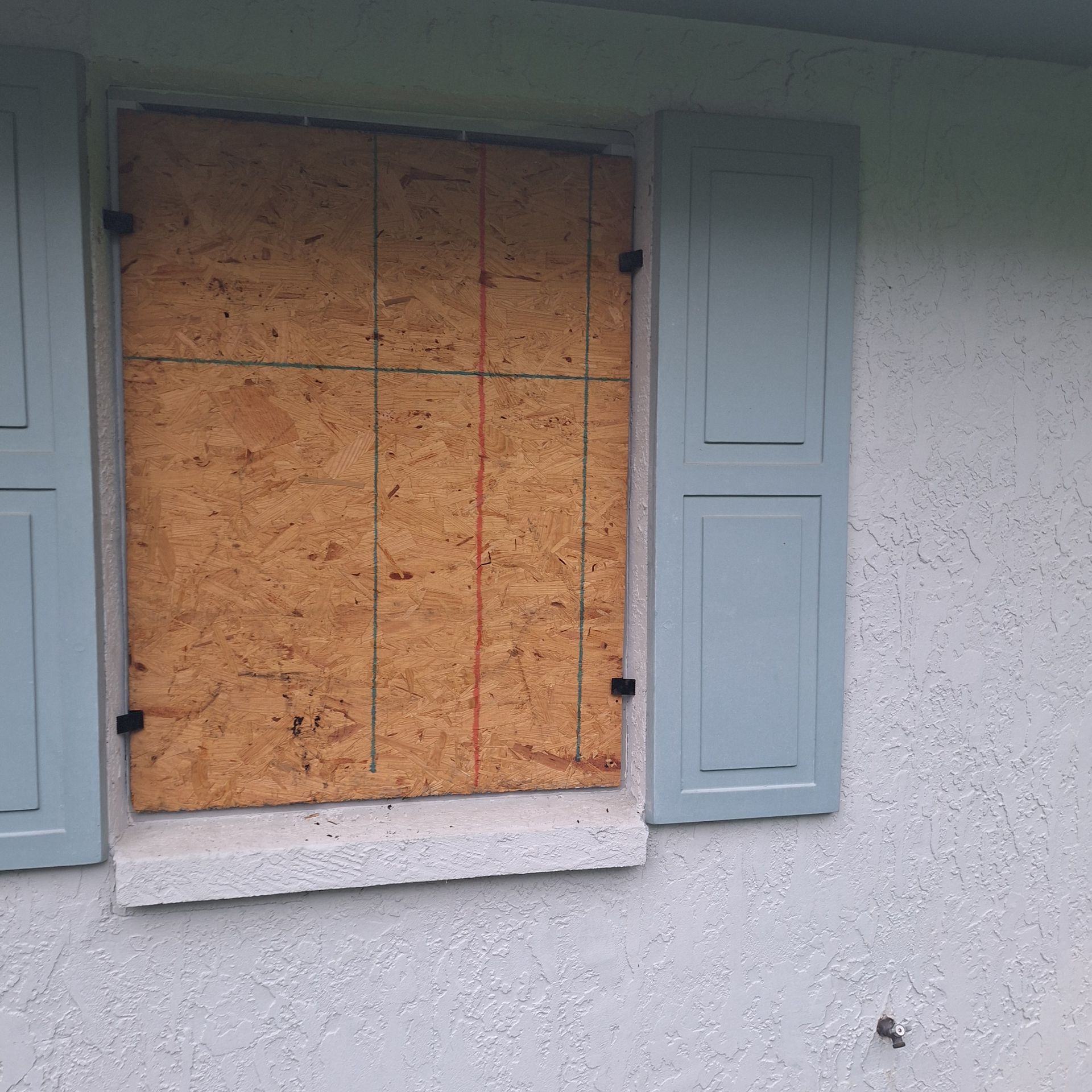 A window with blue shutters is boarded up with plywood