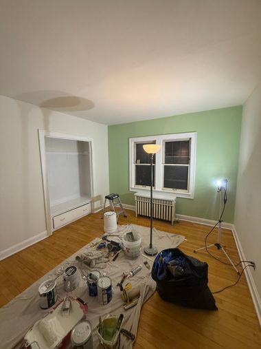 Living room with painting supplies on a tarp, white fireplace, green wall, and window lit by a work lamp.