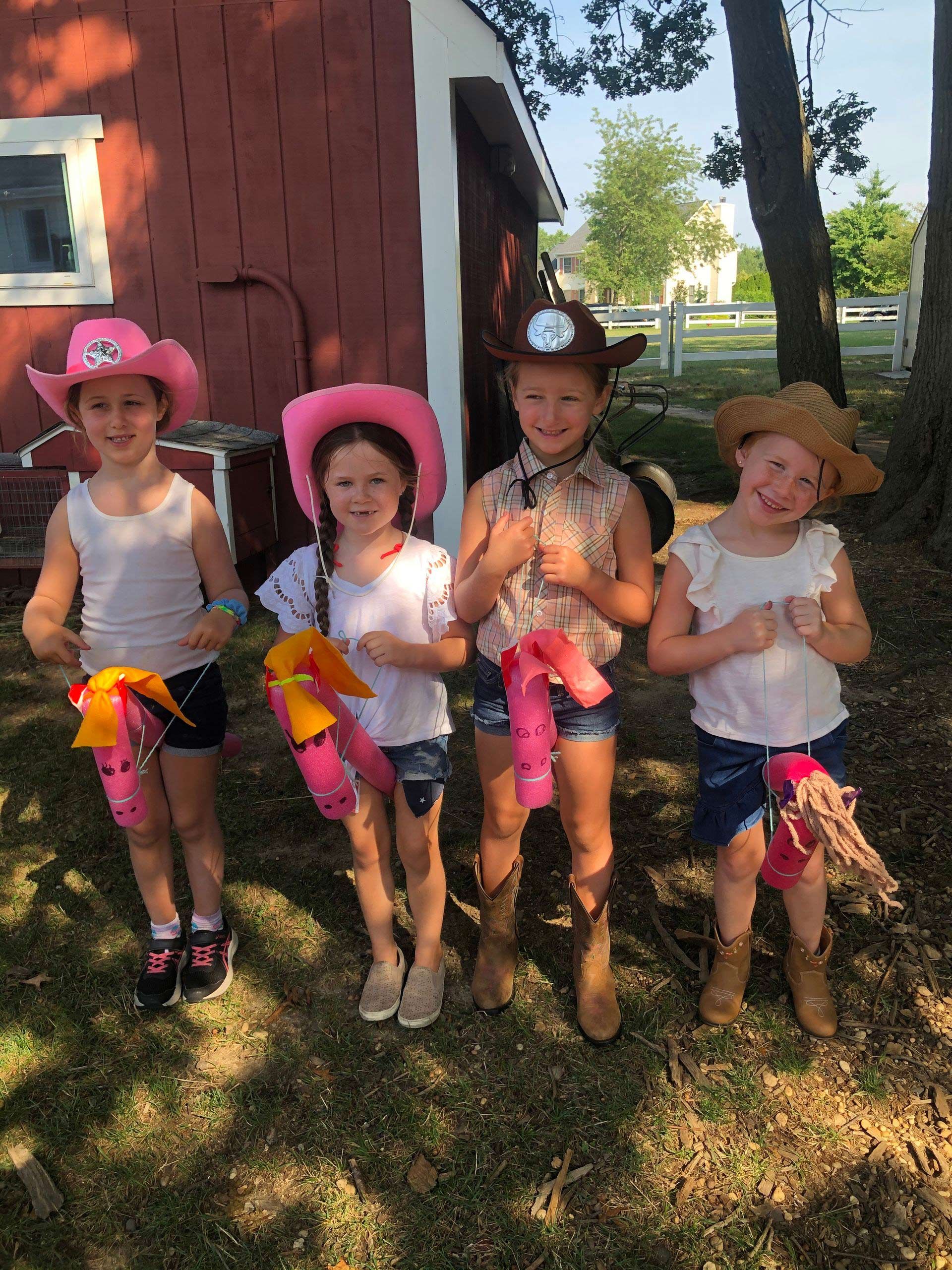 Four little girls wearing cowboy hats and boots are standing in front of a red barn.