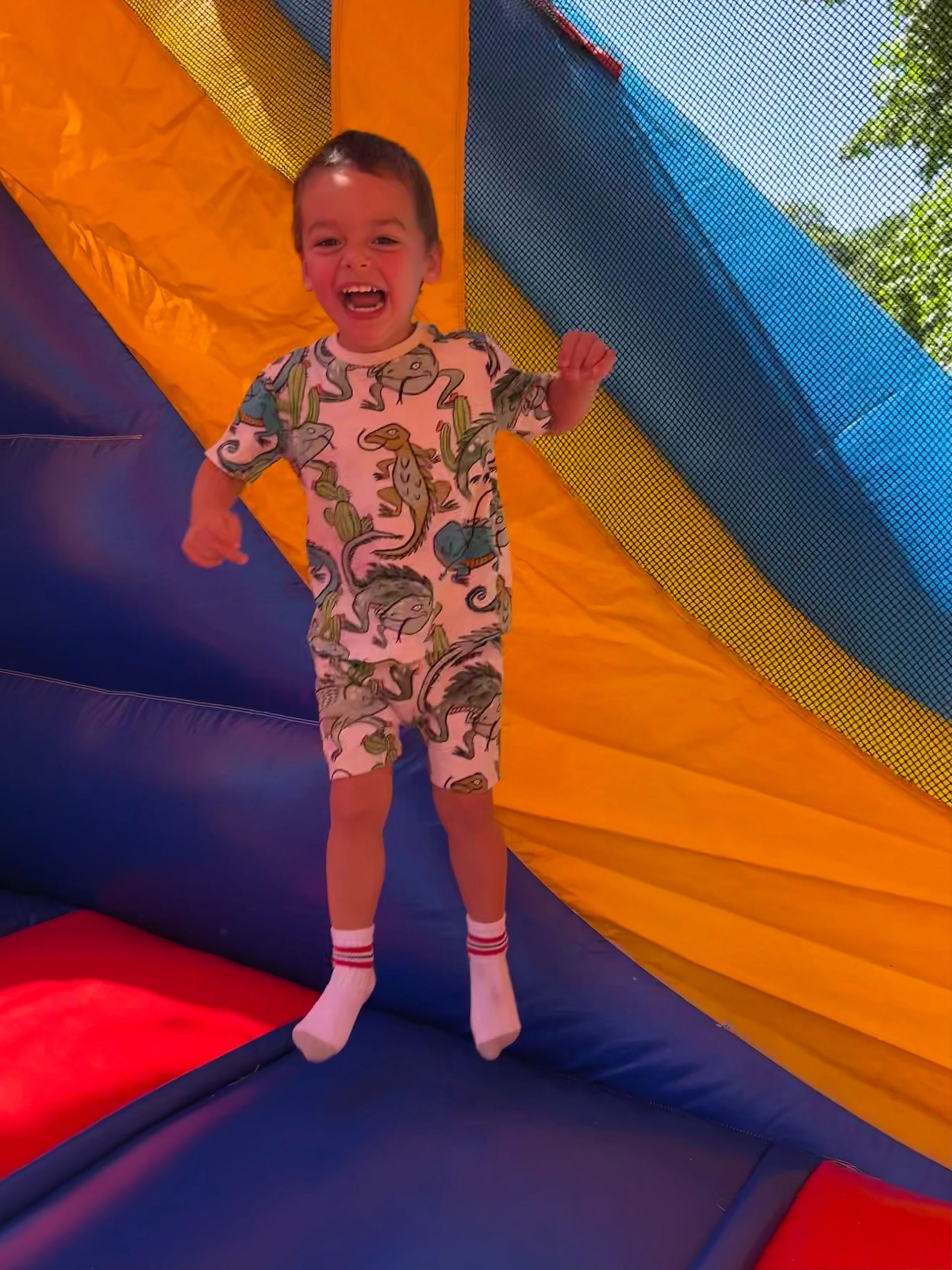 A little boy in a dinosaur shirt is jumping in a bouncy house.