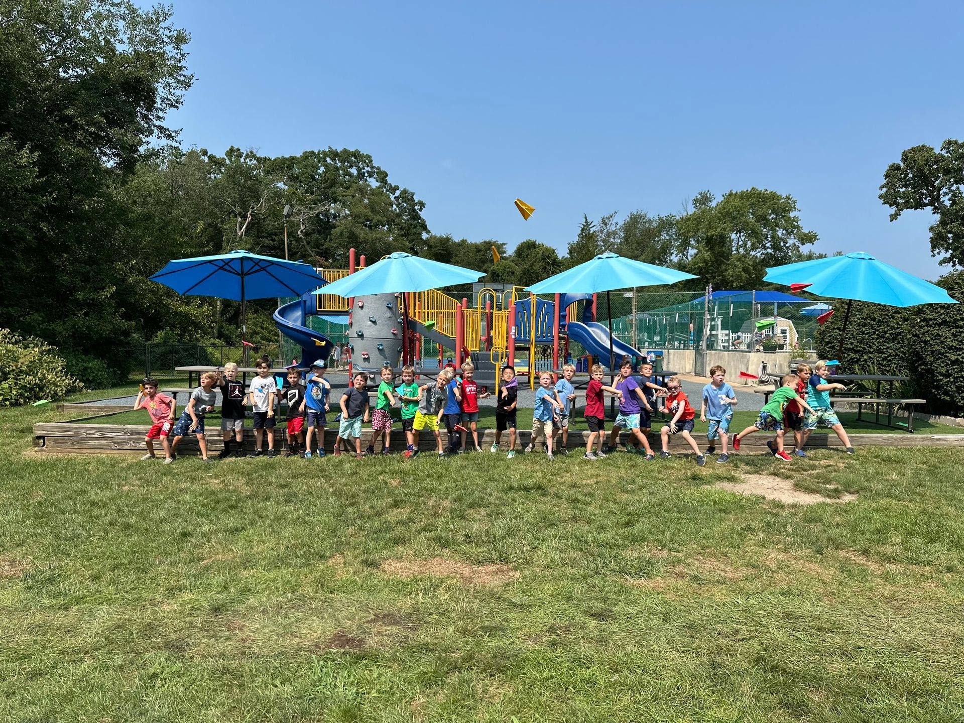 A group of children are standing in a field with umbrellas in front of a playground.