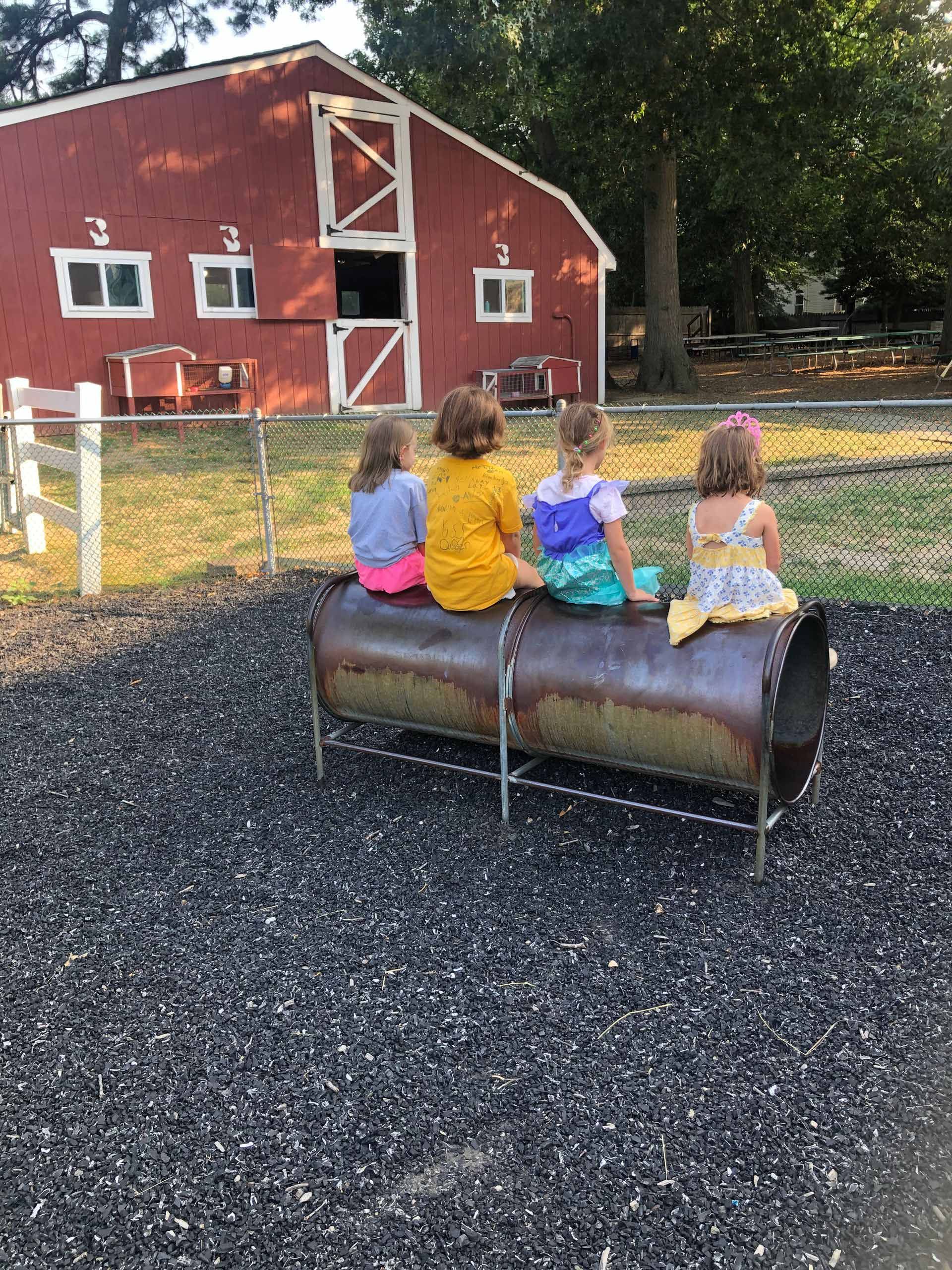 Three little girls are sitting on a pipe in front of a red barn.