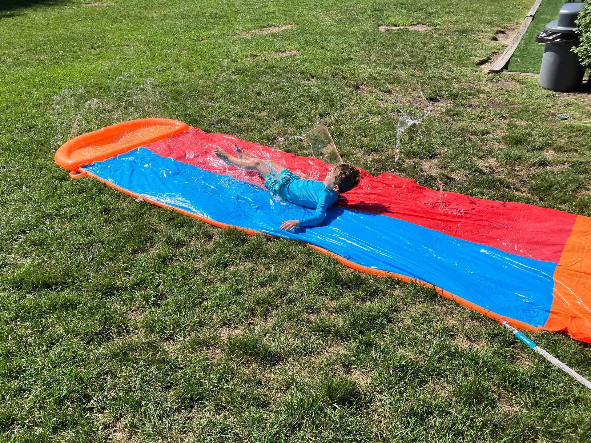 A child is laying on a water slide in the grass.