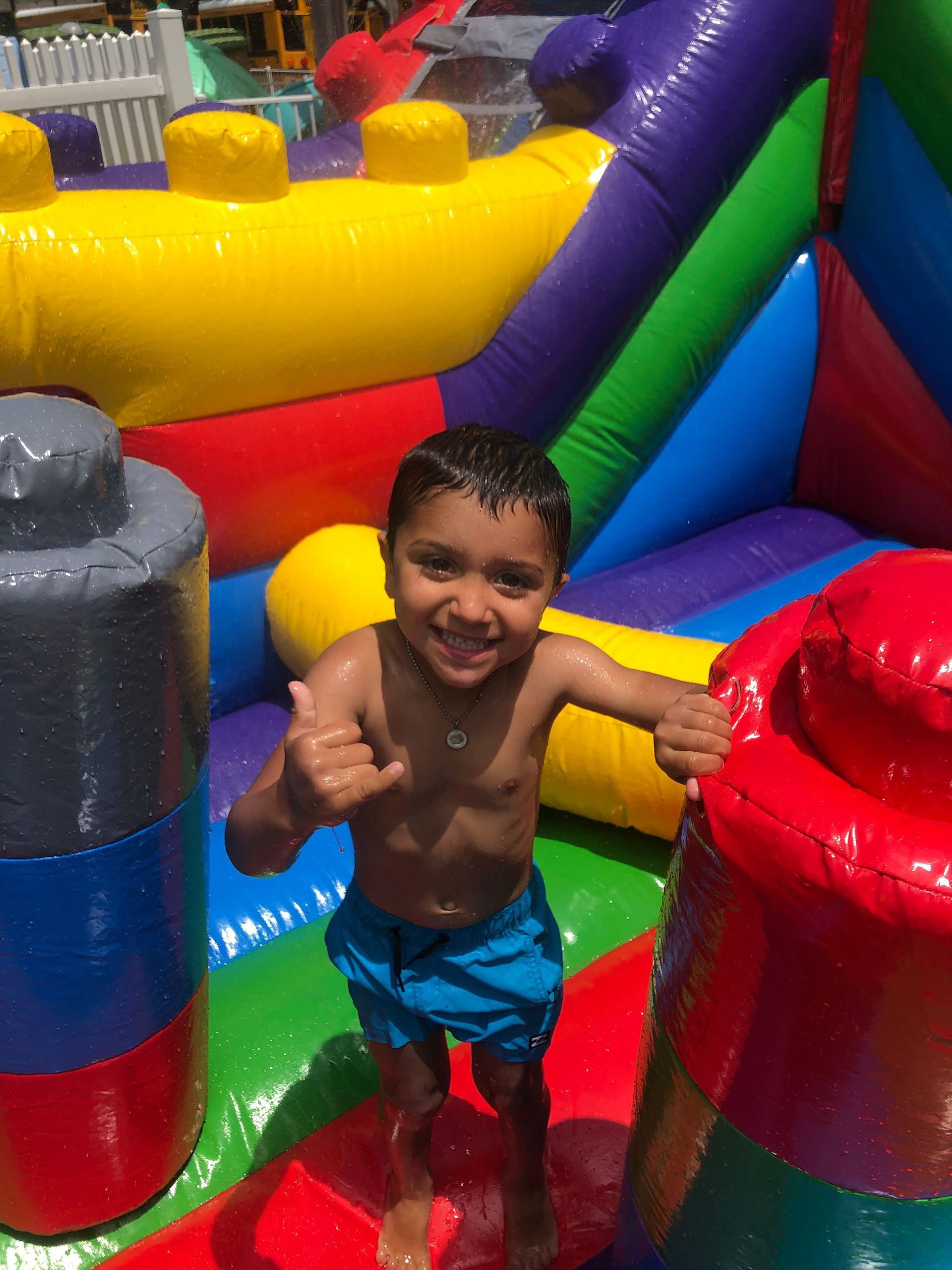 A young boy is standing in front of a colorful bouncy house giving a thumbs up.