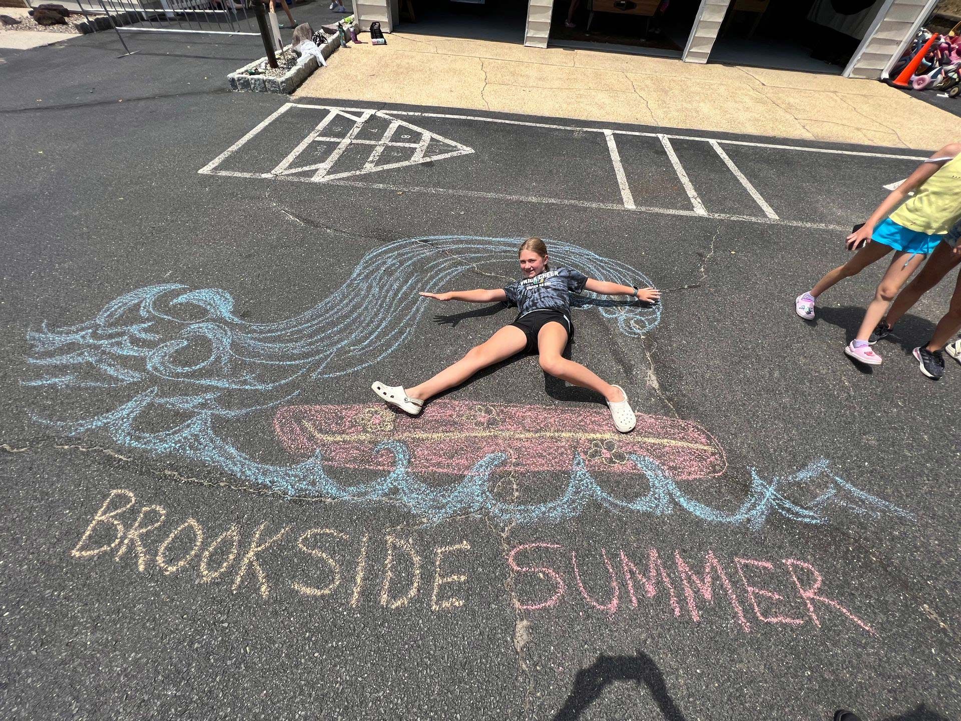 A girl is laying on the ground in front of a chalk drawing of brookside summer.