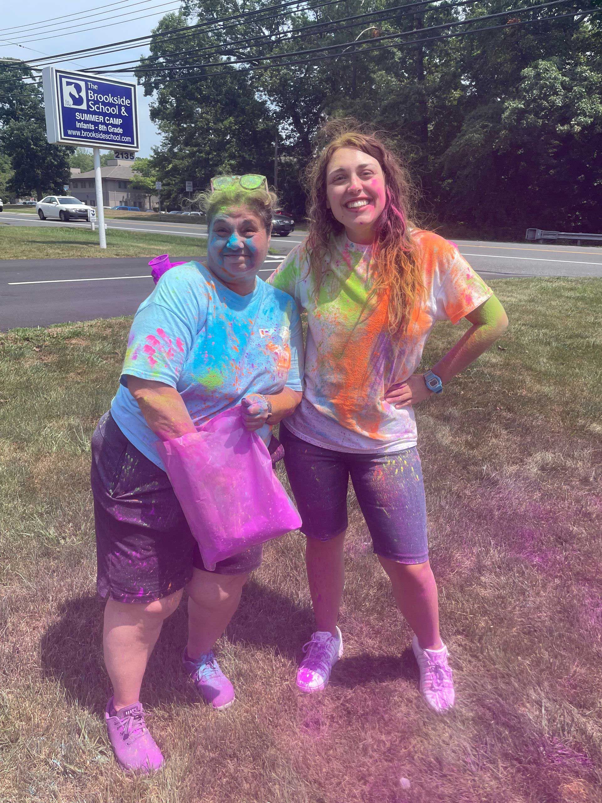 Two women are standing next to each other in the grass covered in colored powder.