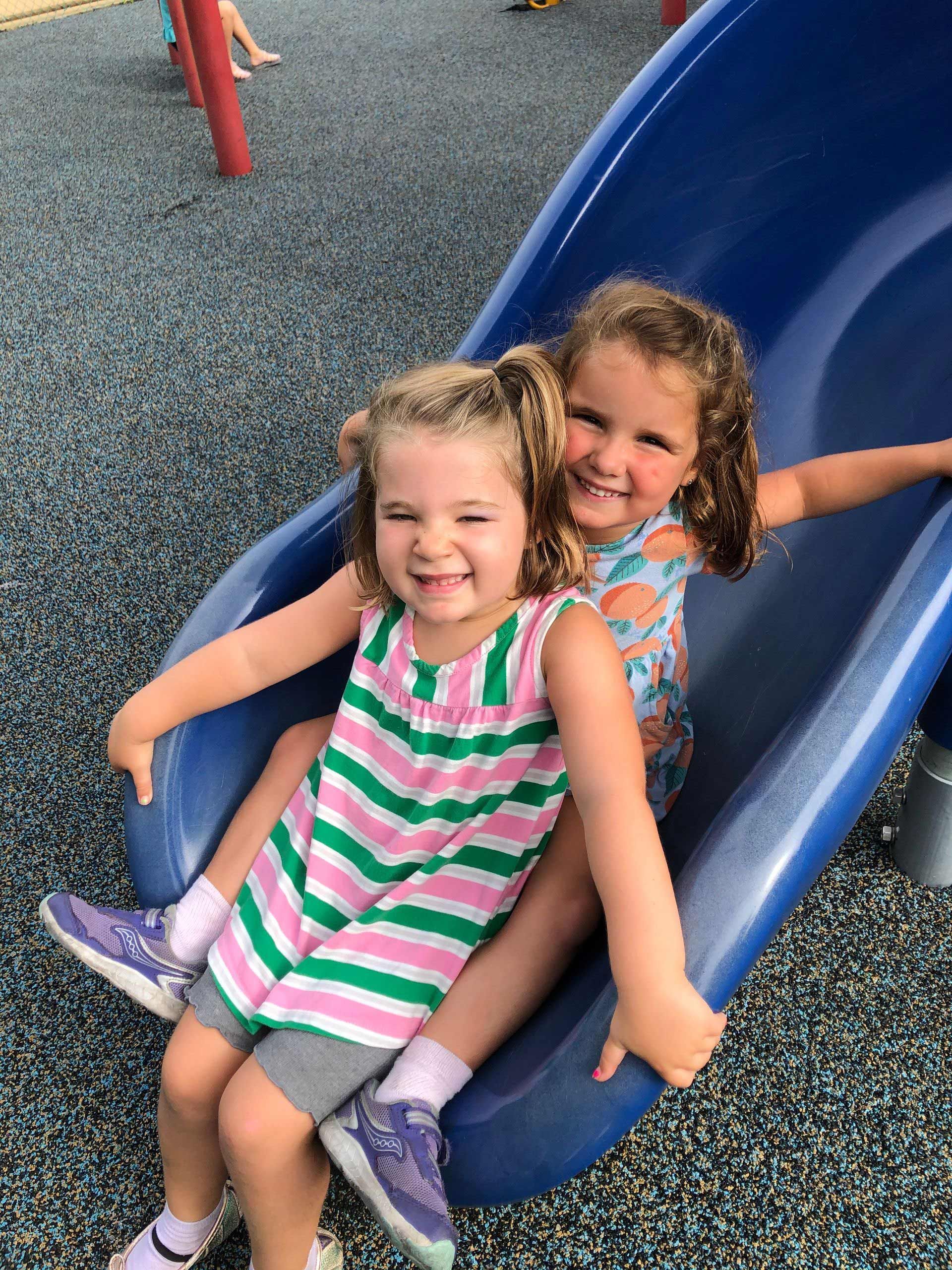 Two little girls are sitting on a slide at a playground.