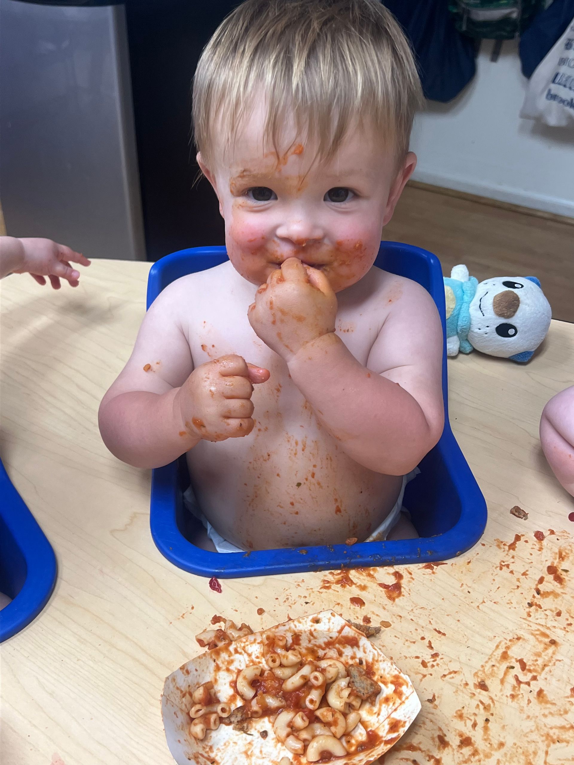 A baby is sitting in a blue bowl eating macaroni and cheese.