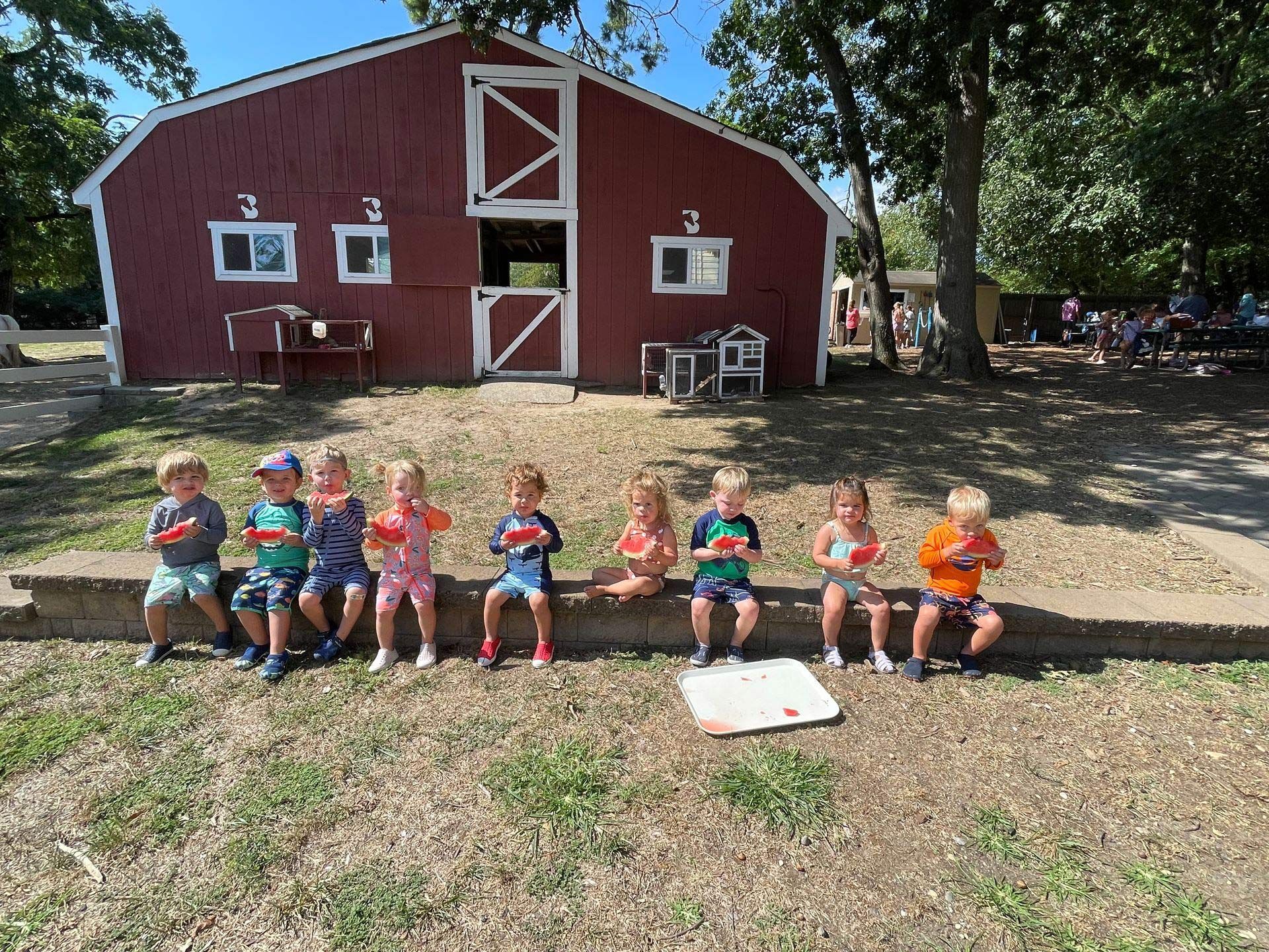 A group of children are sitting on a bench in front of a barn eating watermelon.