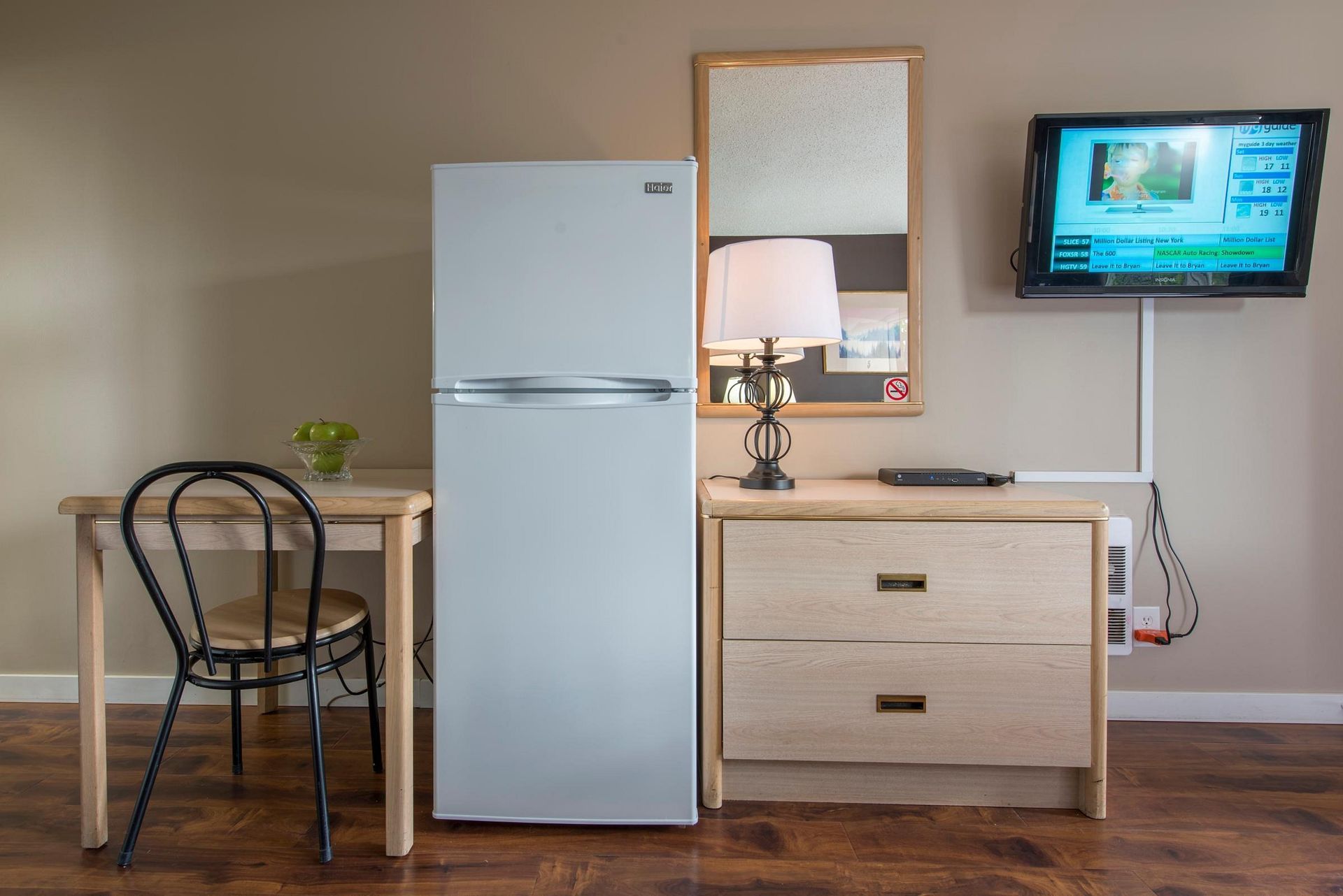 A room with a refrigerator , dresser , table and television.