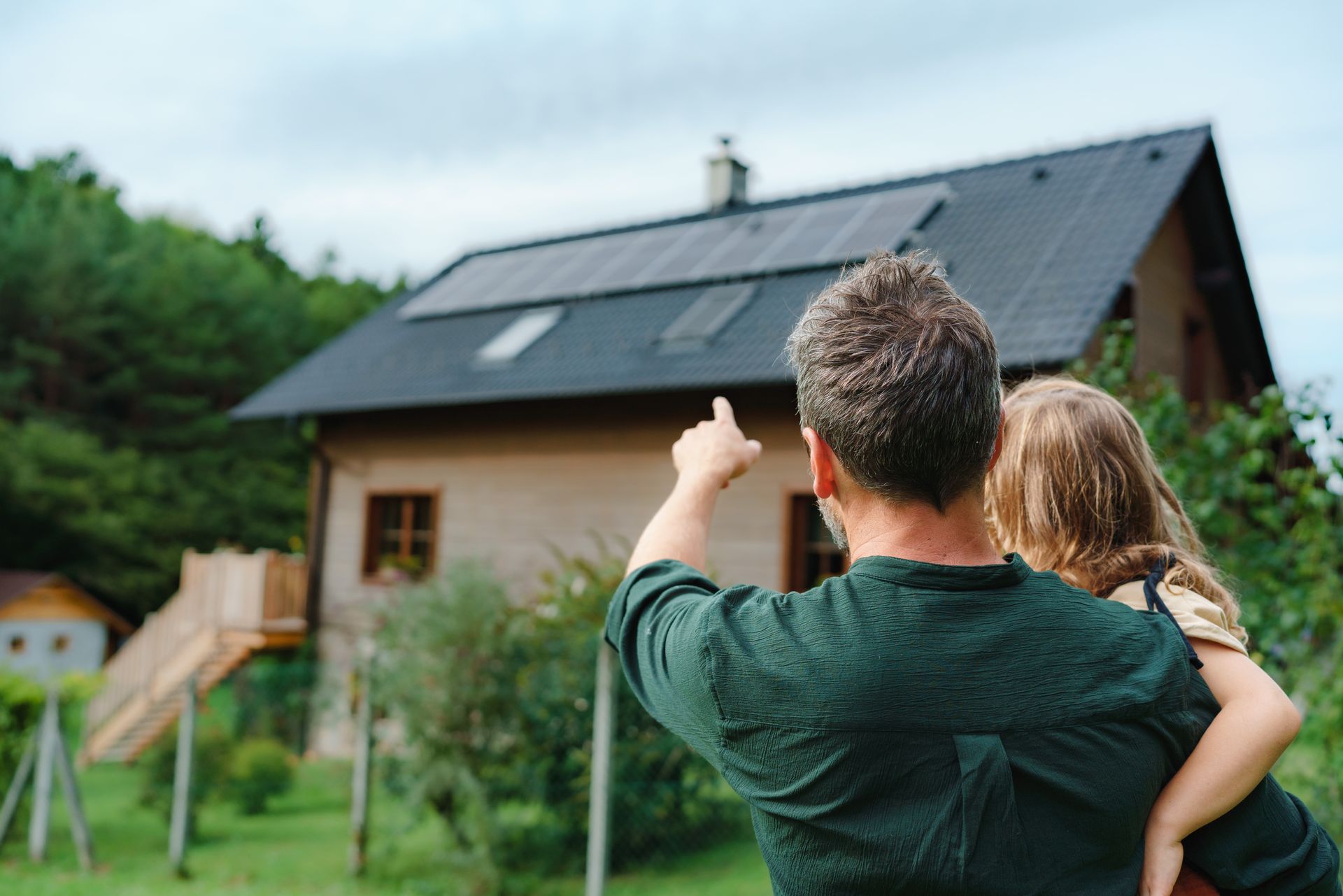 Man points at solar panels on a house roof while holding a child; green yard, trees, cloudy sky.
