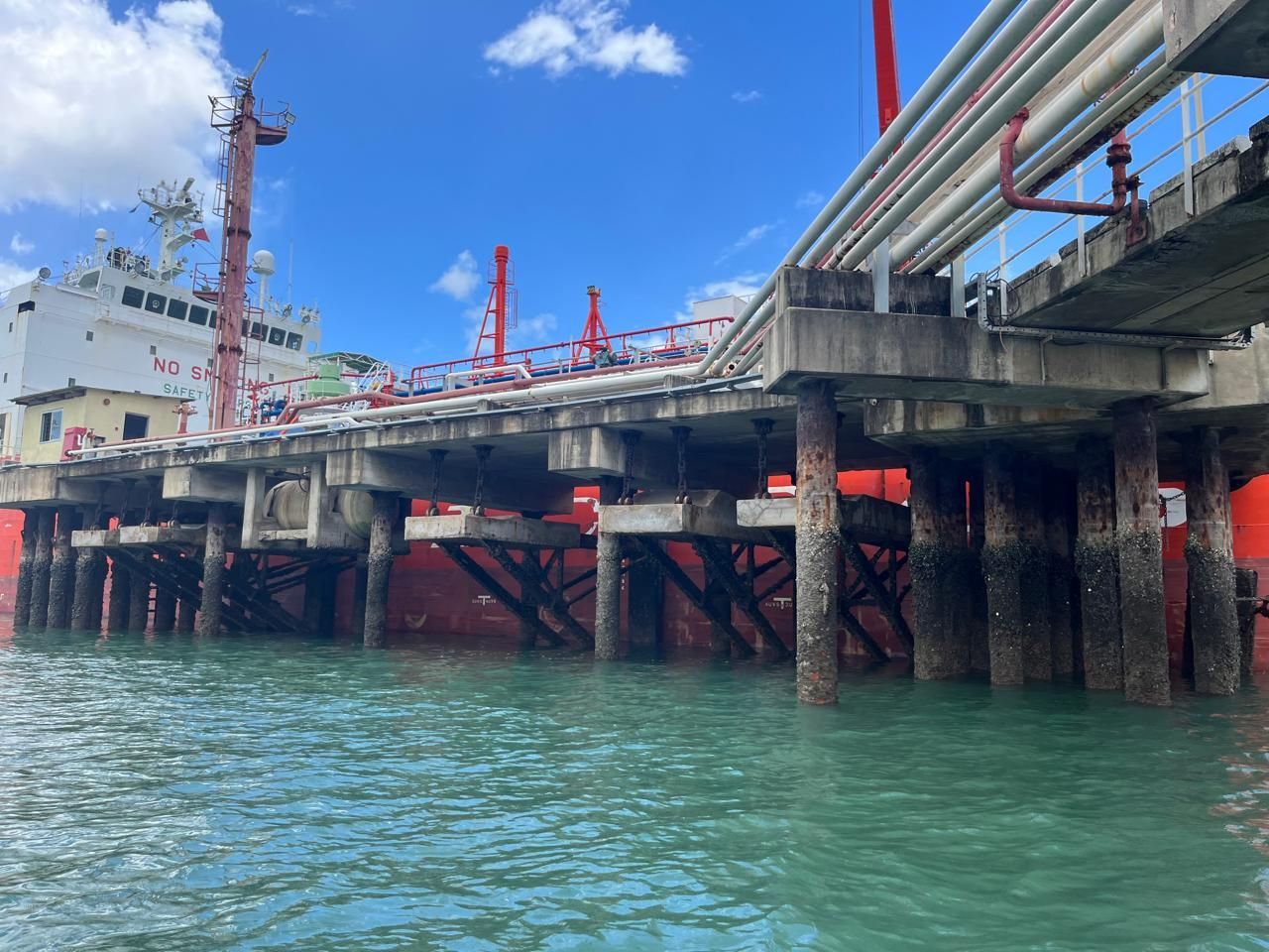A red ship docked at a pier with pipes, supported by wooden pilings in blue-green water under a blue sky.