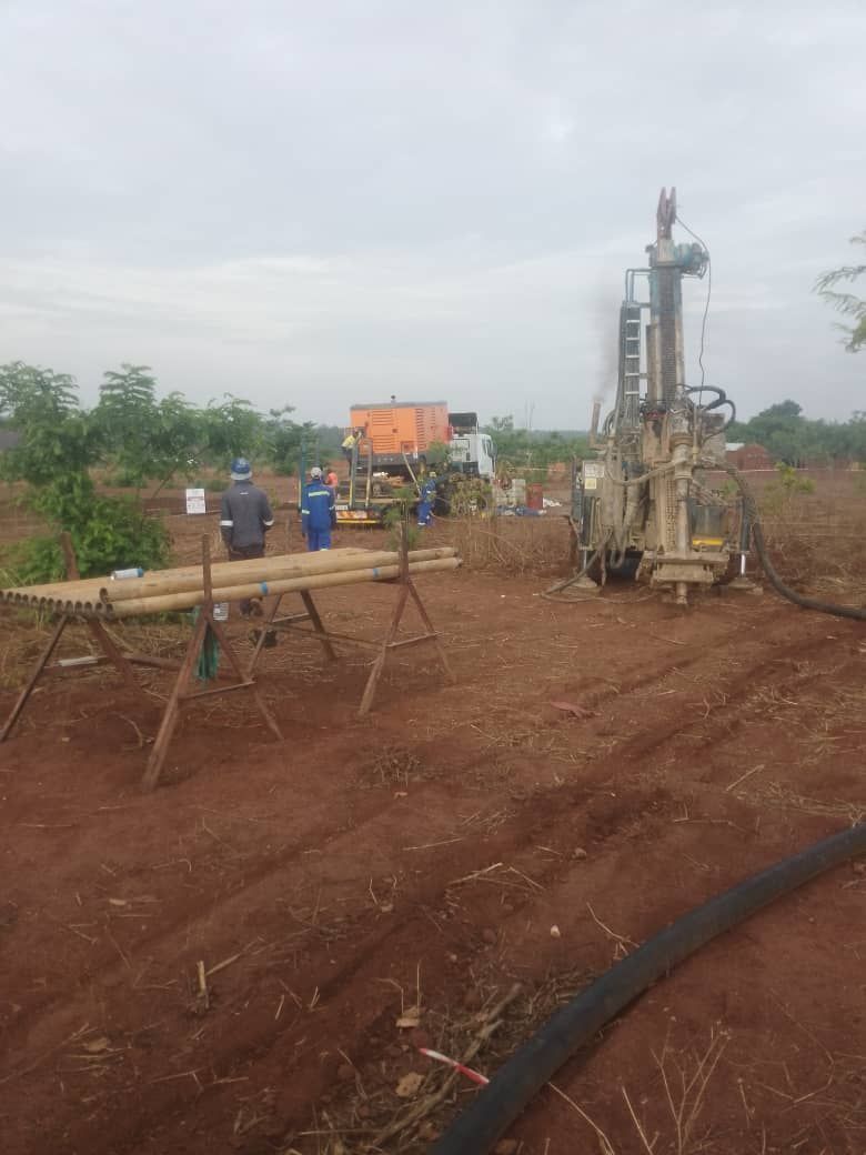 Drilling rig in a field; two workers in blue overalls near a support structure with pipes; cloudy sky.