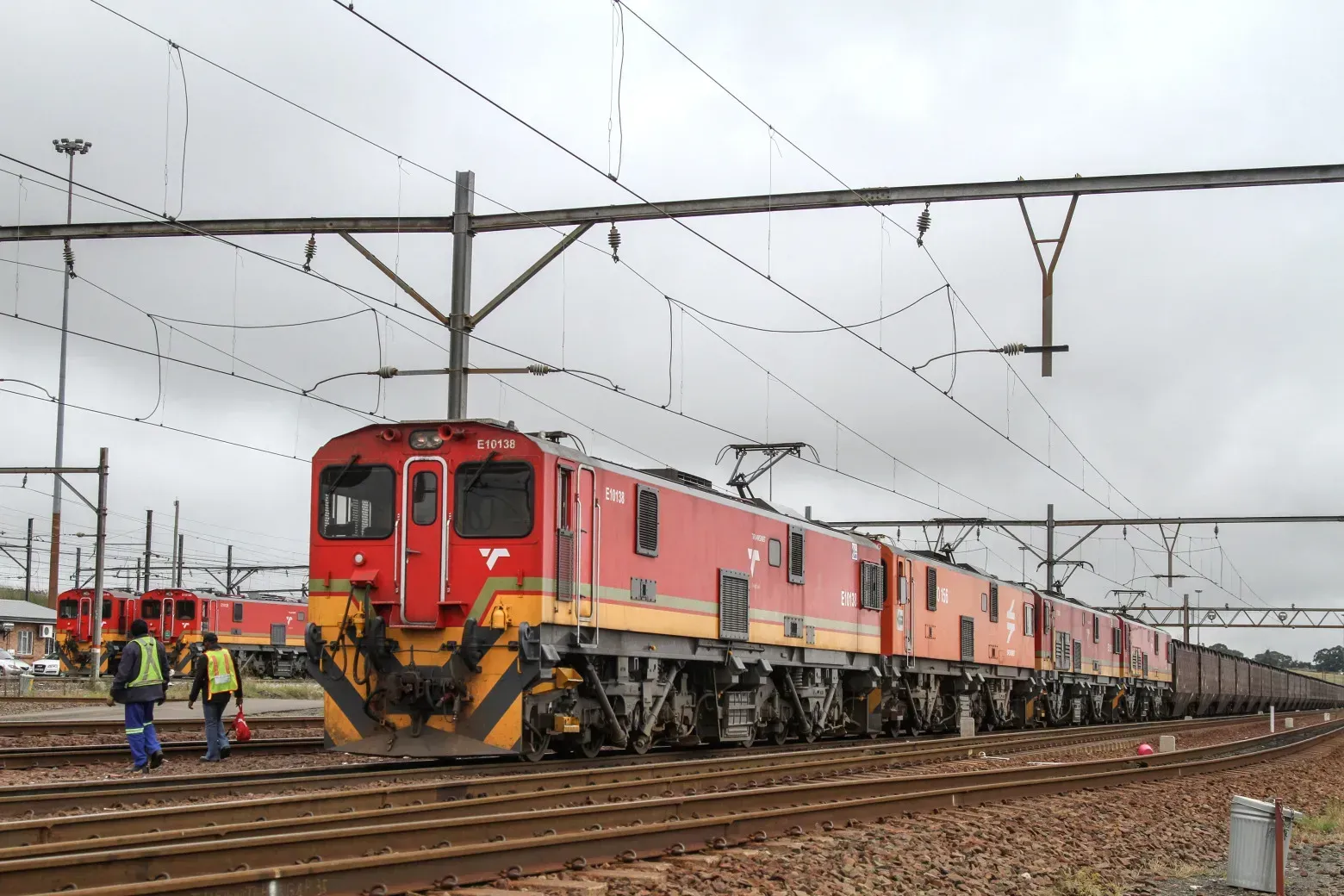 Red and yellow electric train on tracks, under overhead wires. Two figures near the train.