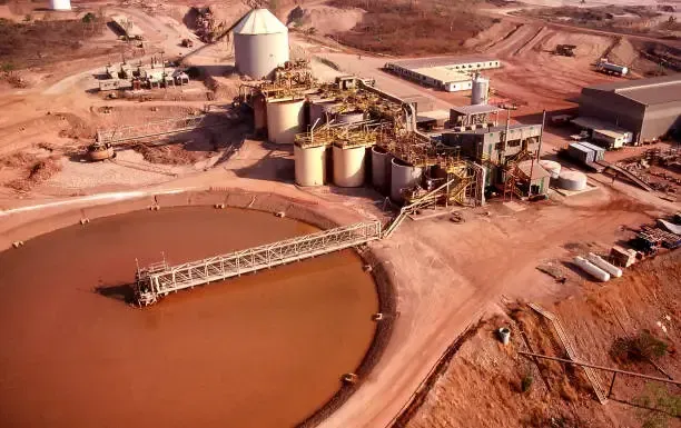 Aerial view of a brown mining facility with tanks, buildings, and a large waste pond with a walkway.