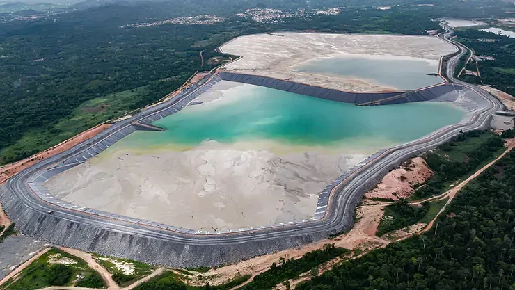 Large industrial tailings pond with gray liner, light blue-green water, and surrounding forest.