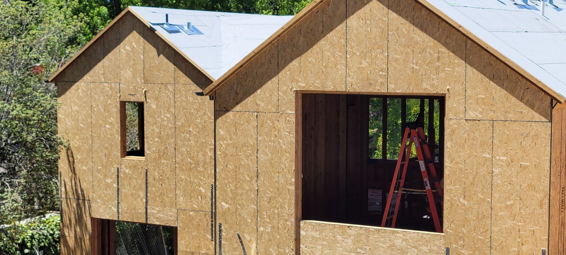 Exterior view of a house under construction with oriented strand board siding and a ladder inside a window.