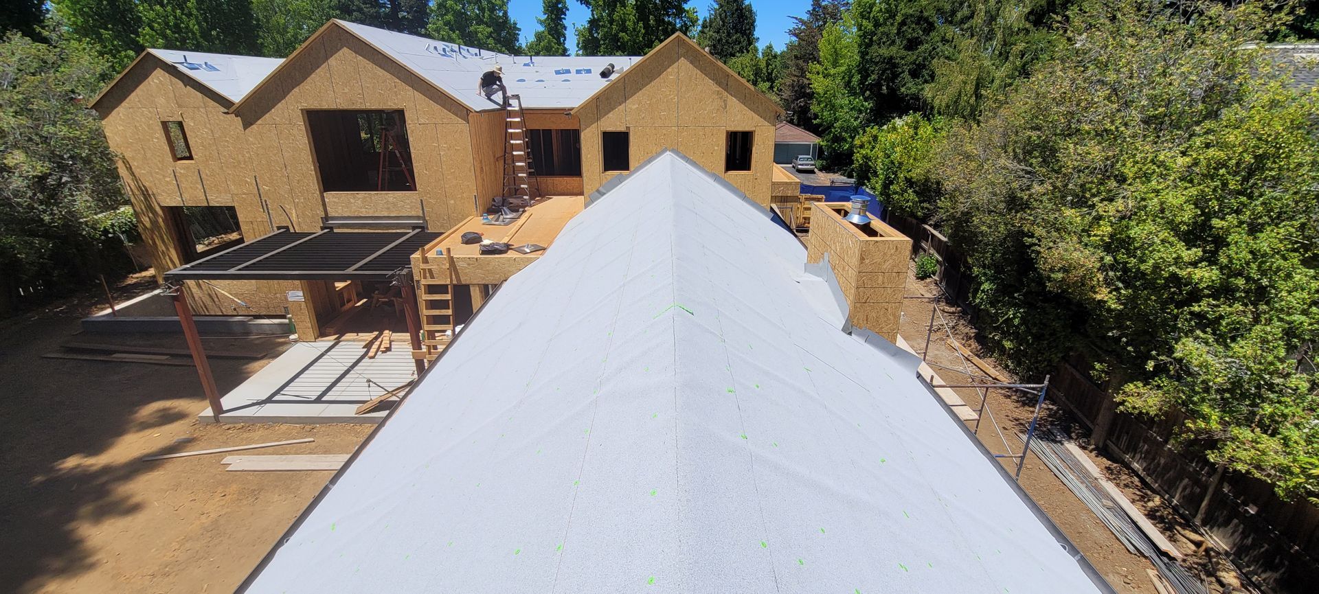 A house under construction with a large white roof and wooden frame, set amongst trees.