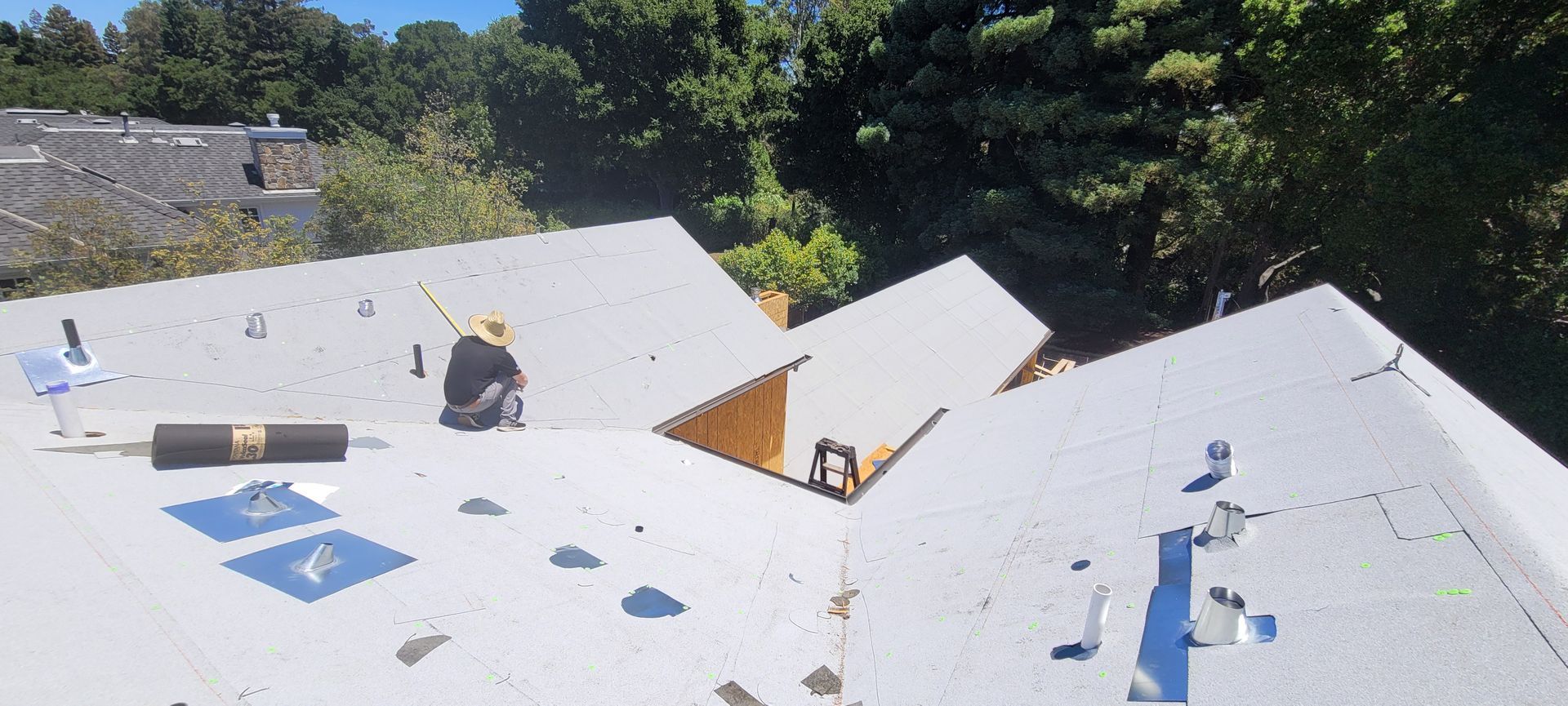 A person works on a white metal roof with trees and blue sky in the background.