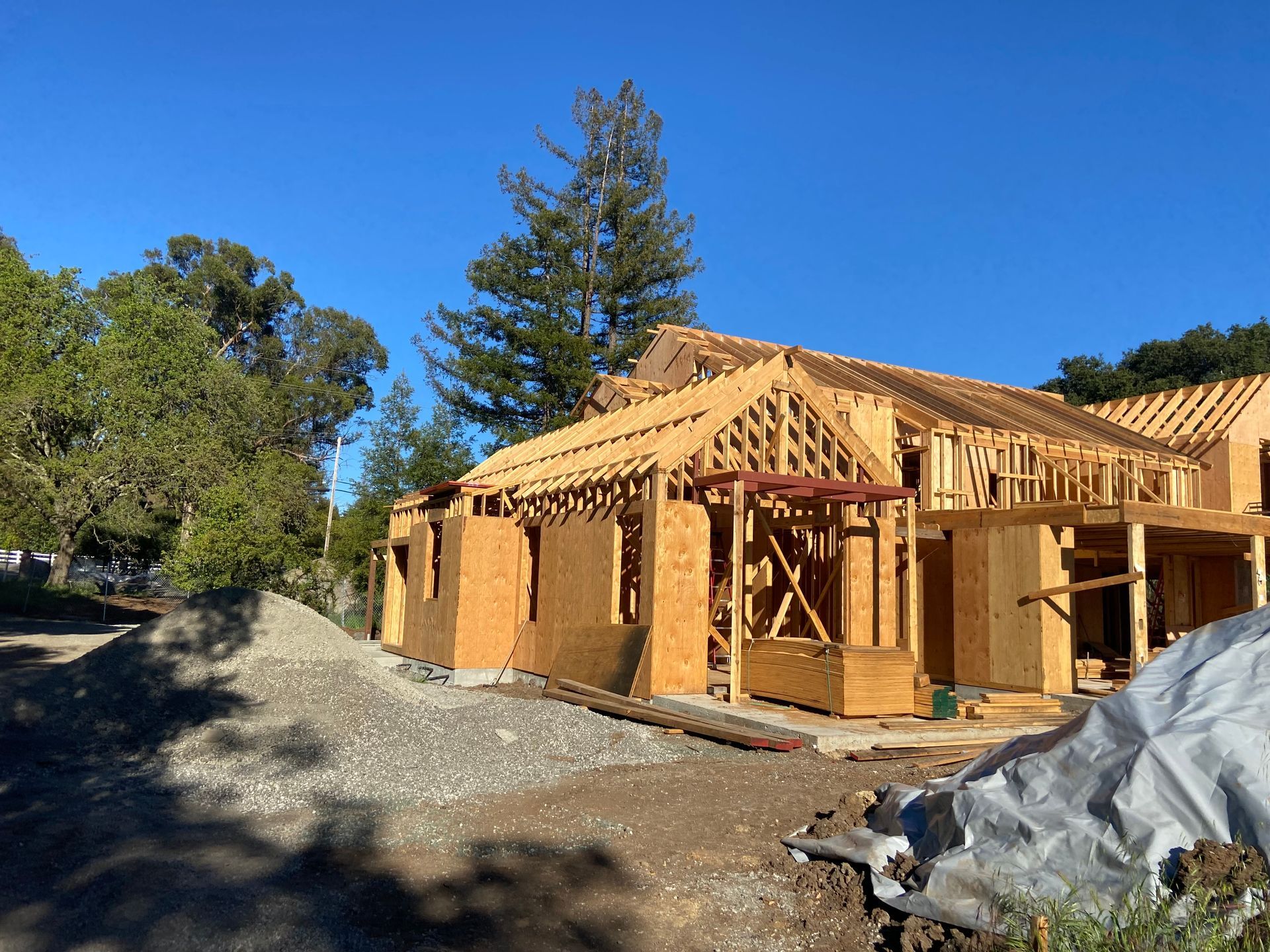 Construction of a wood-framed house under a blue sky, with surrounding trees and gravel.
