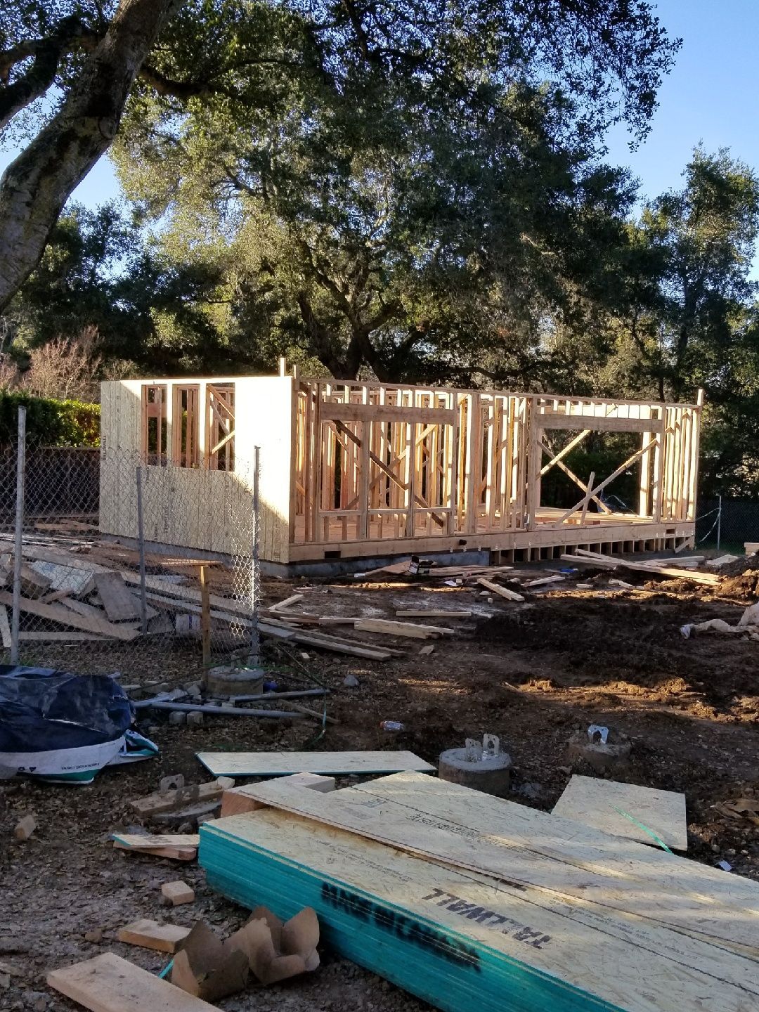 Wooden house frame under construction outdoors. Lumber, plywood, and dirt visible.