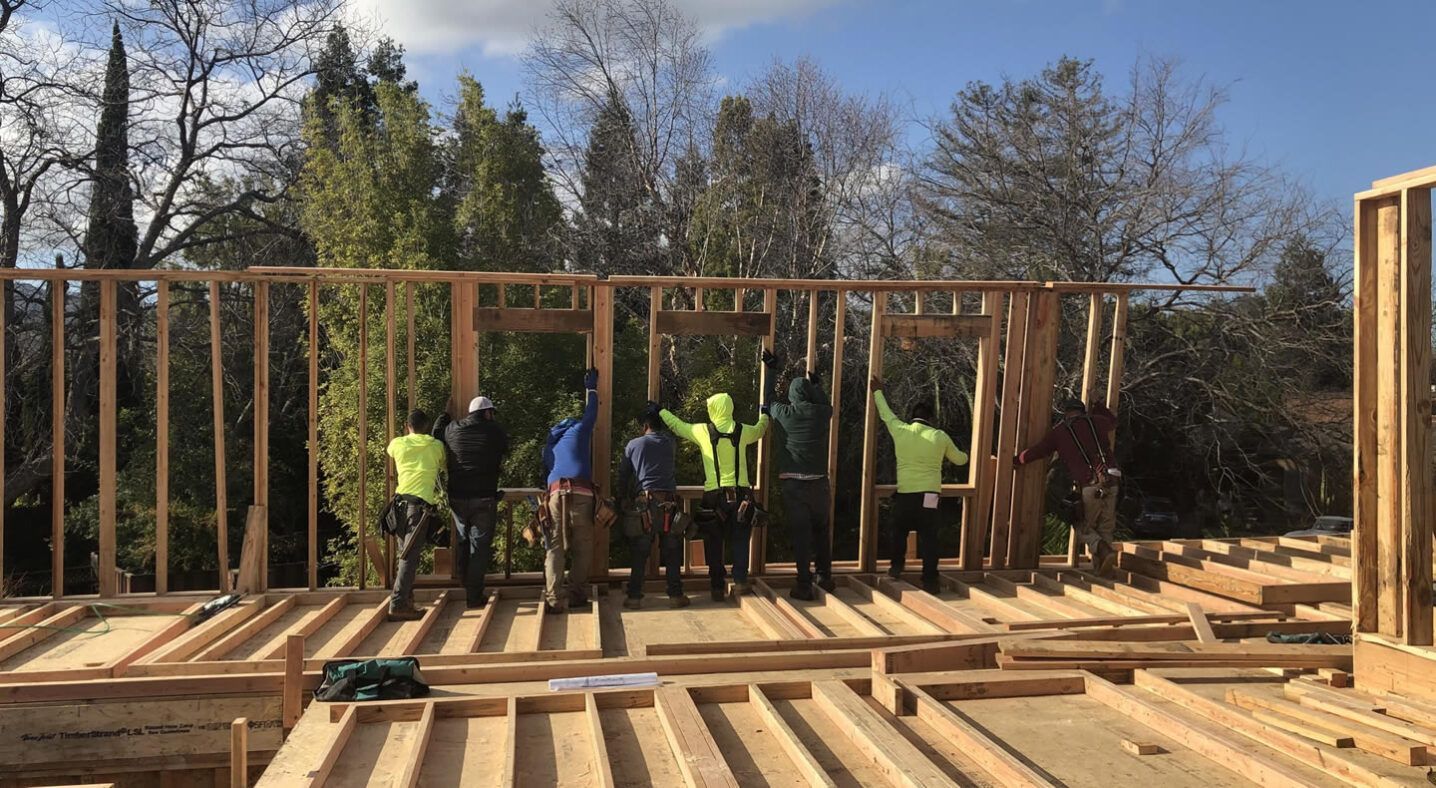 Construction workers raising a wooden wall frame on a building site, with trees in the background.