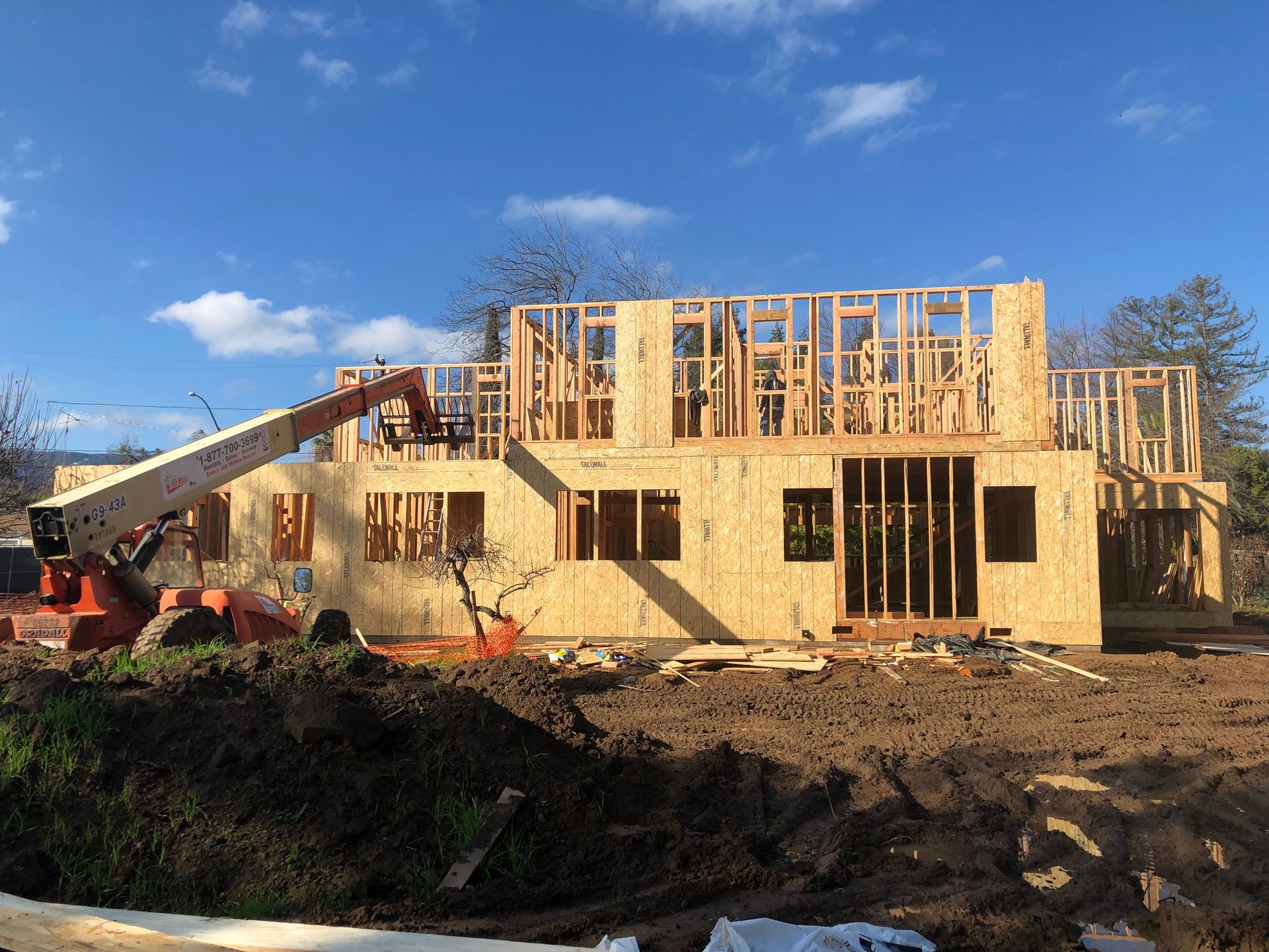 House under construction; exposed wood roof, white protective wrap, garage, blue sky, trees.