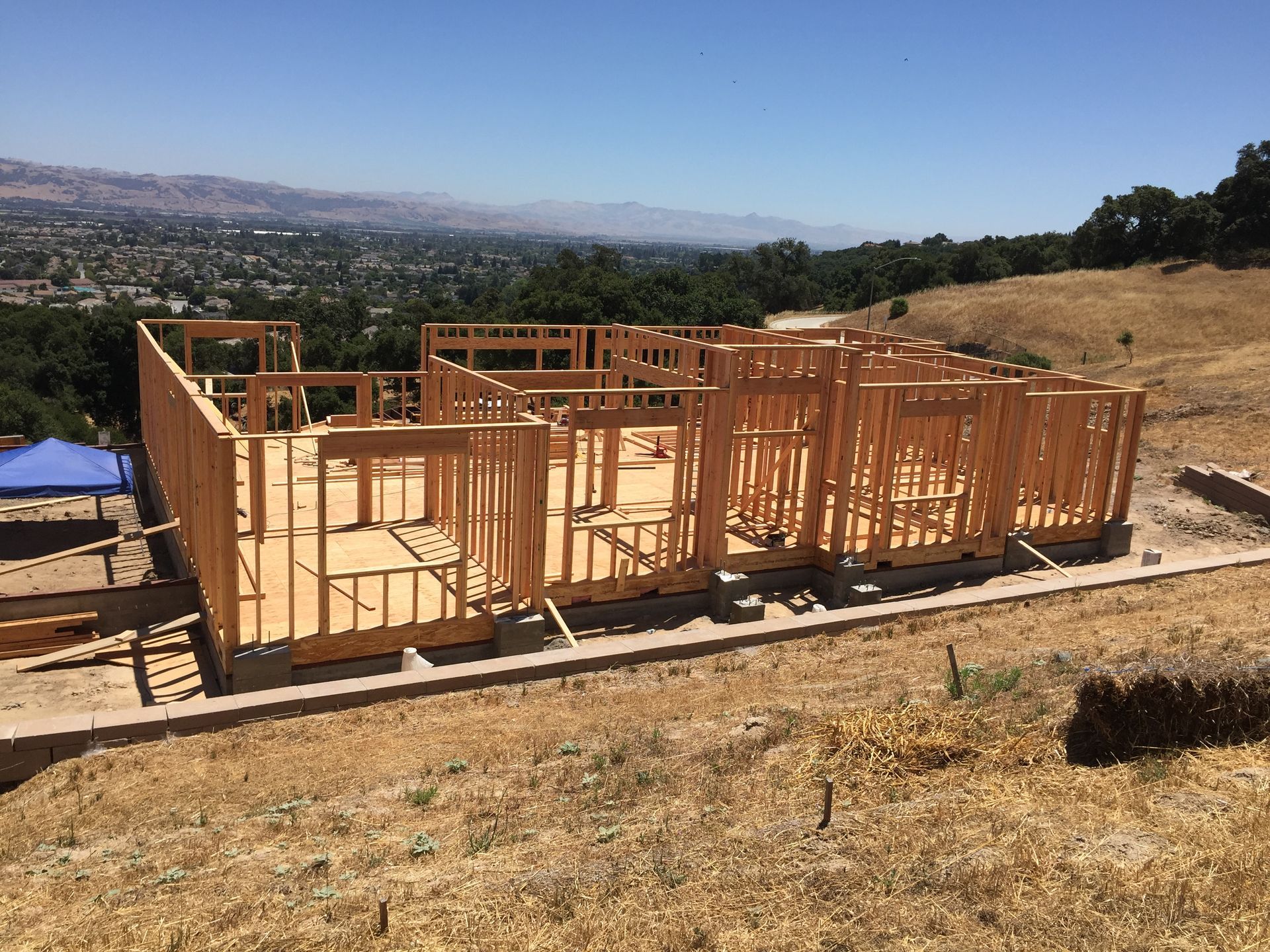 Wooden frame of a house under construction on a hillside, with distant mountains and blue sky.
