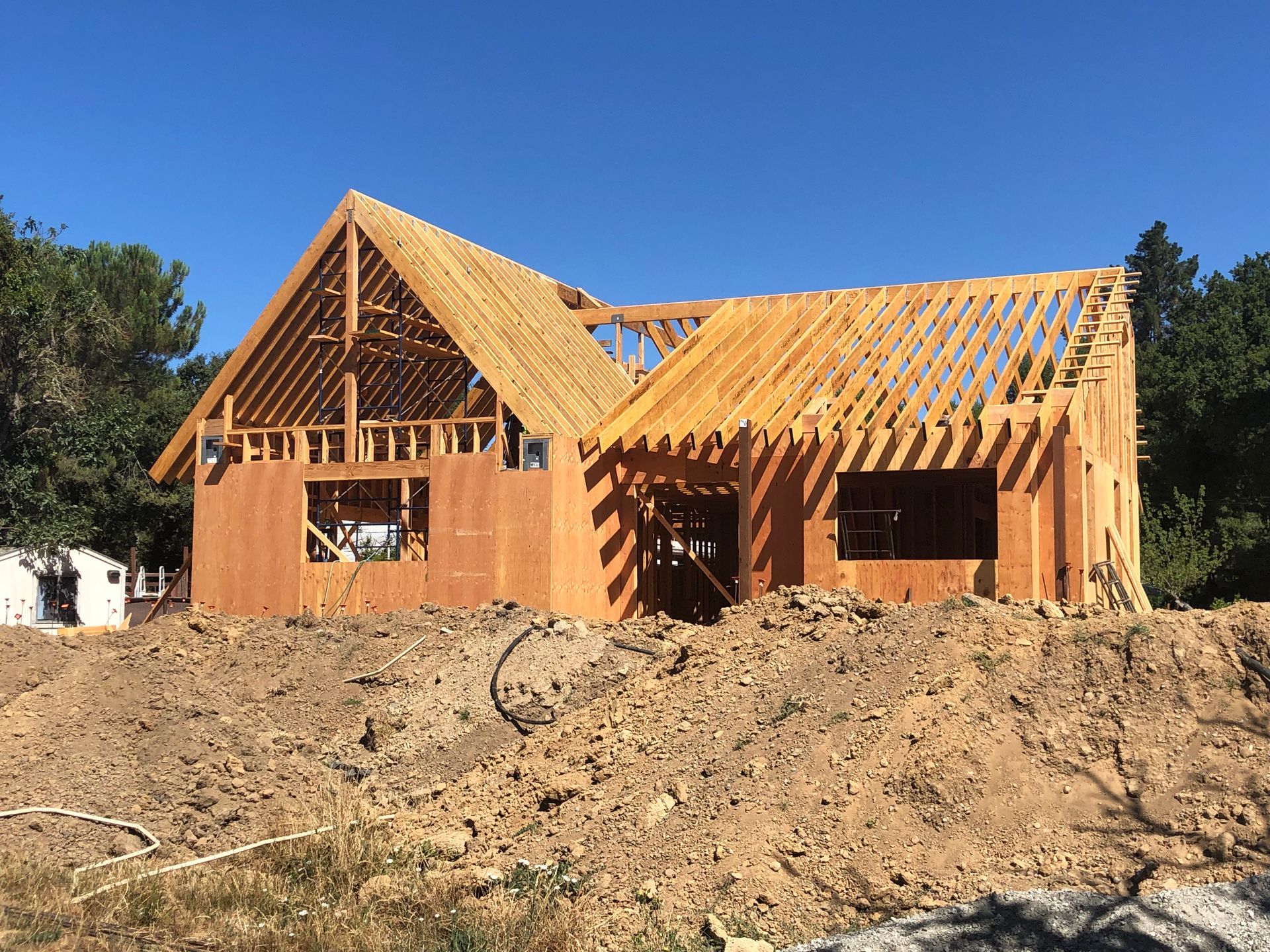 House under construction; wooden frame and roof trusses on dirt, blue sky.