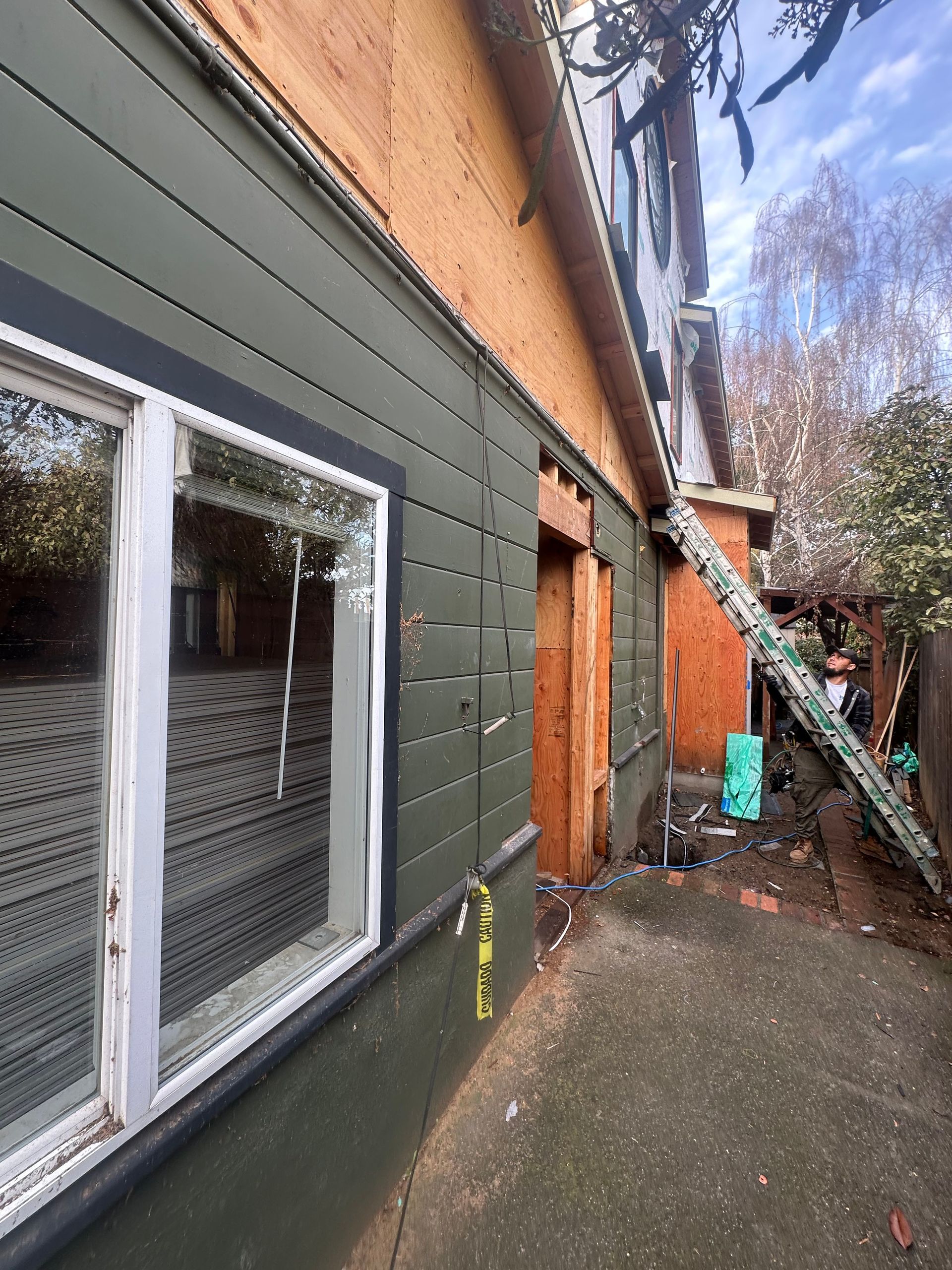 Exterior view of a house under renovation. Green siding, exposed wood, ladder, and a window are visible.