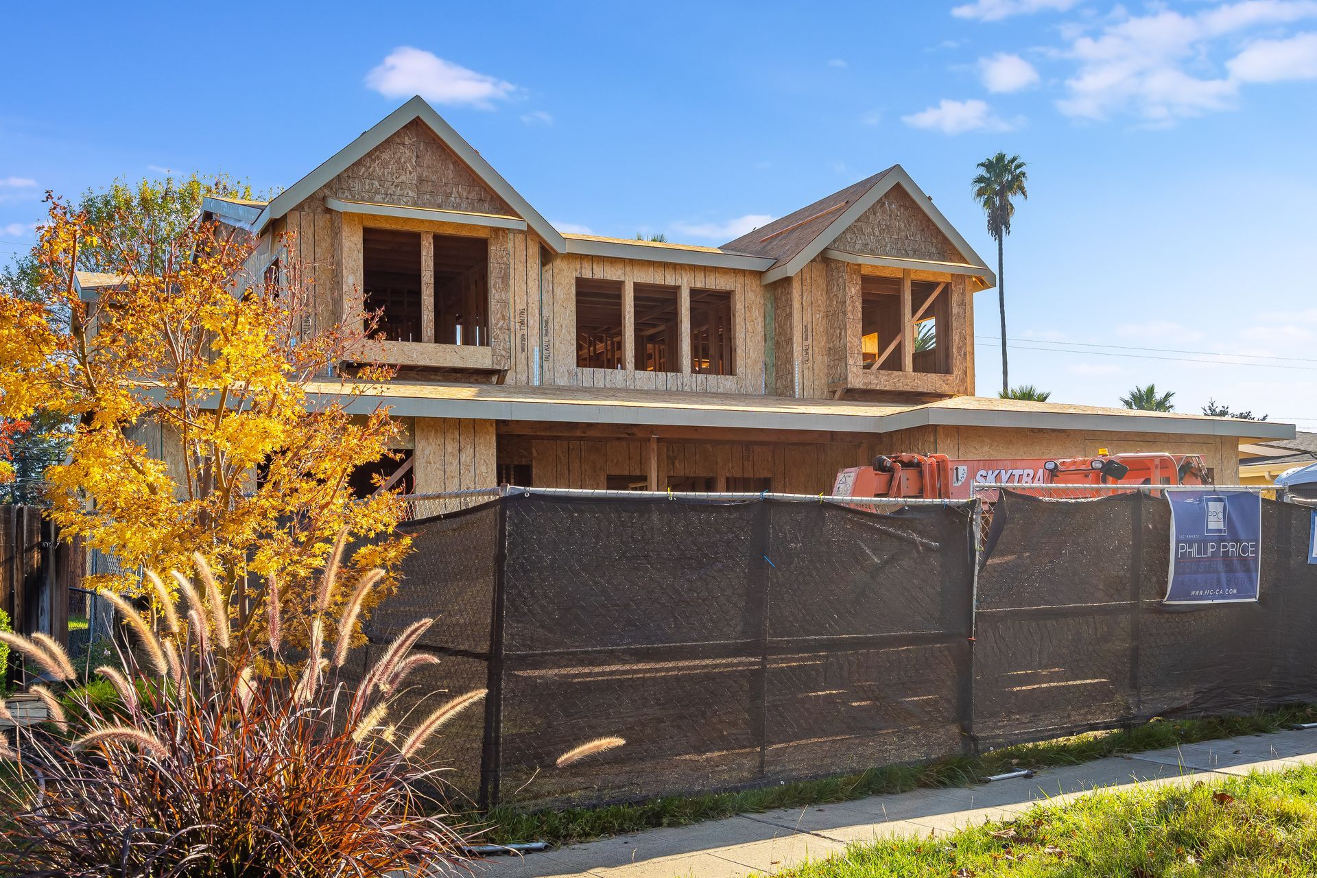 Construction of a two-story house with wood framing, behind a black construction fence on a sunny day.