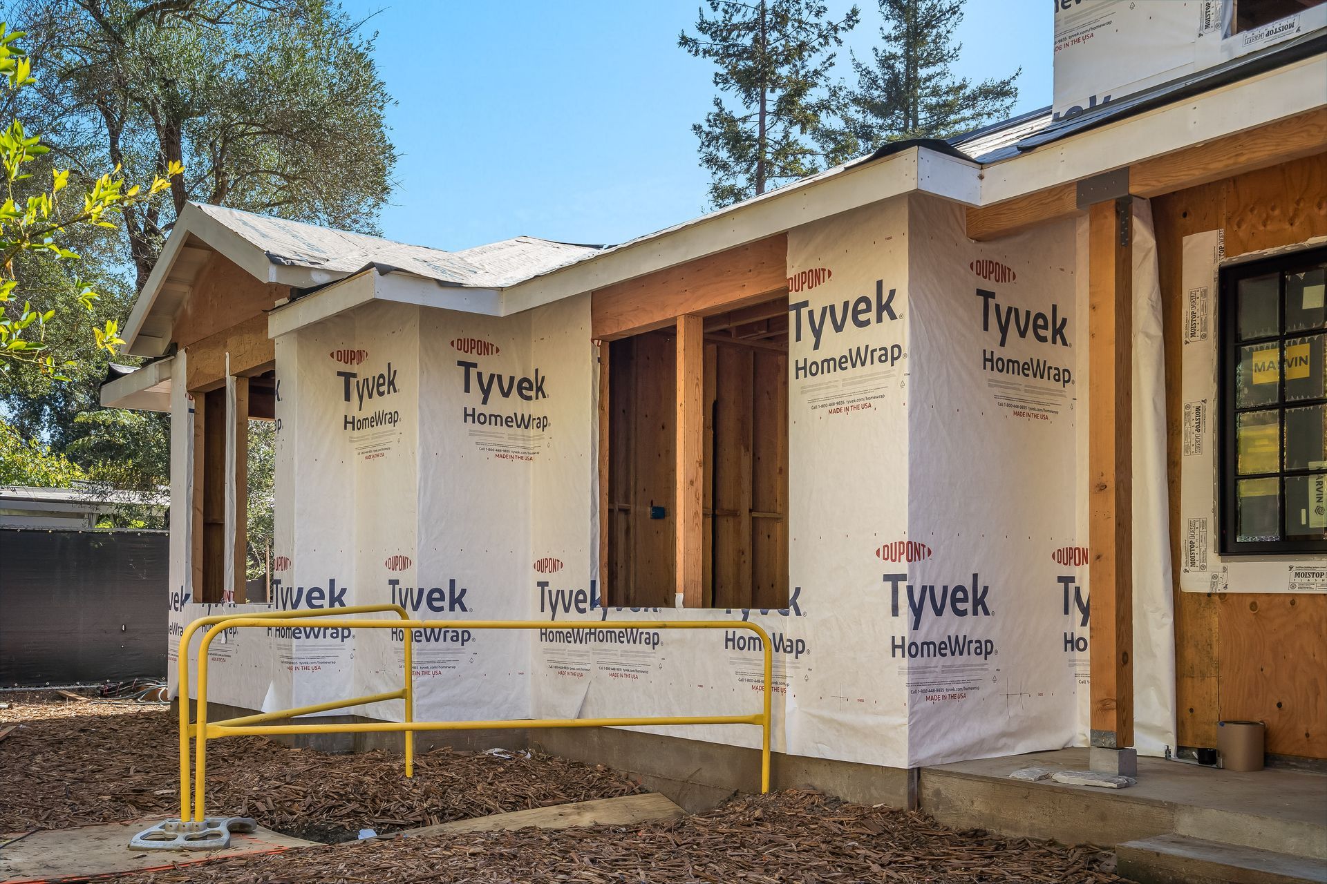 Construction site: building exterior with Tyvek wrap, wooden framing, unfinished openings, and a yellow safety barrier.