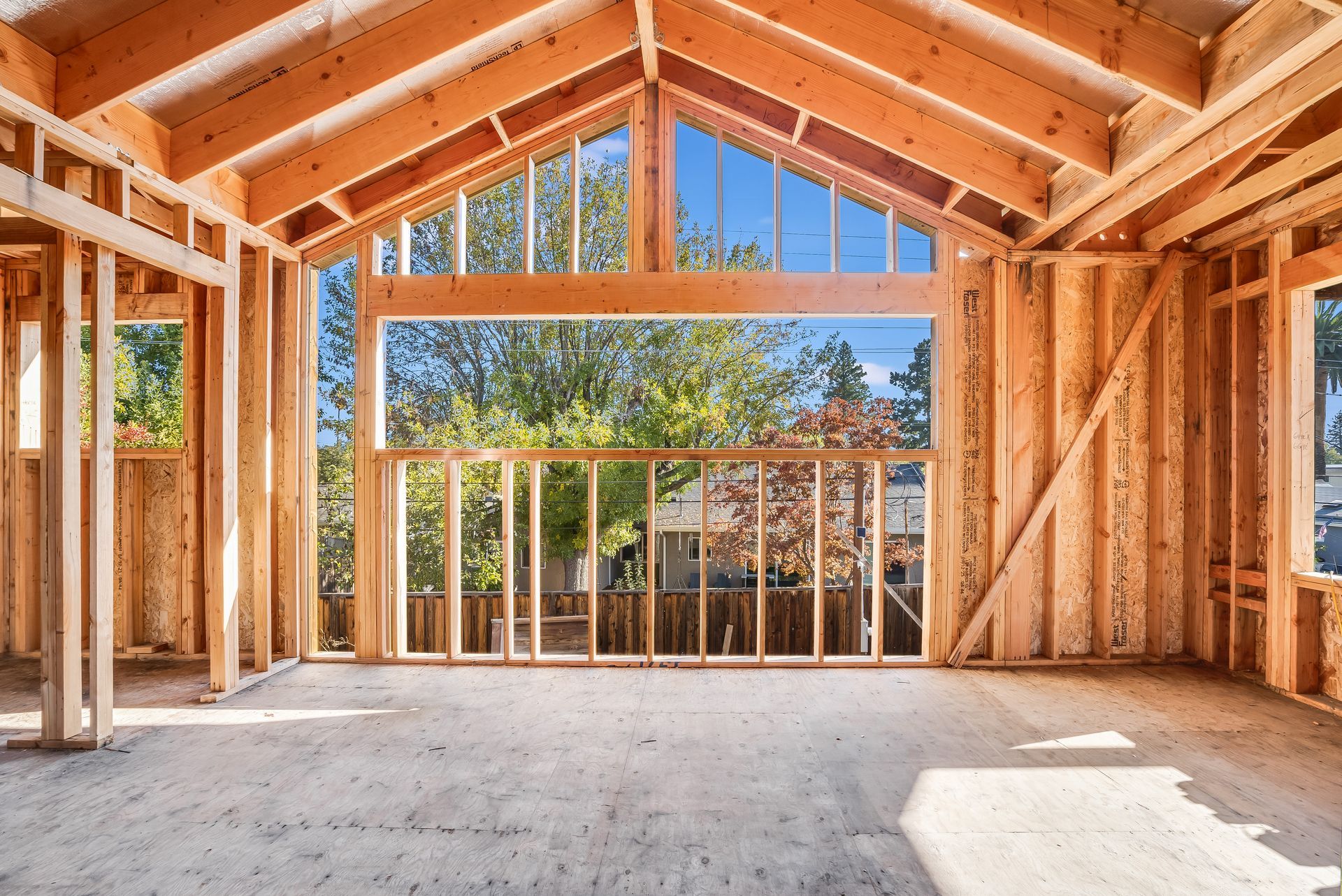 Interior of a house under construction with exposed wooden framing and large windows overlooking a tree-lined area.